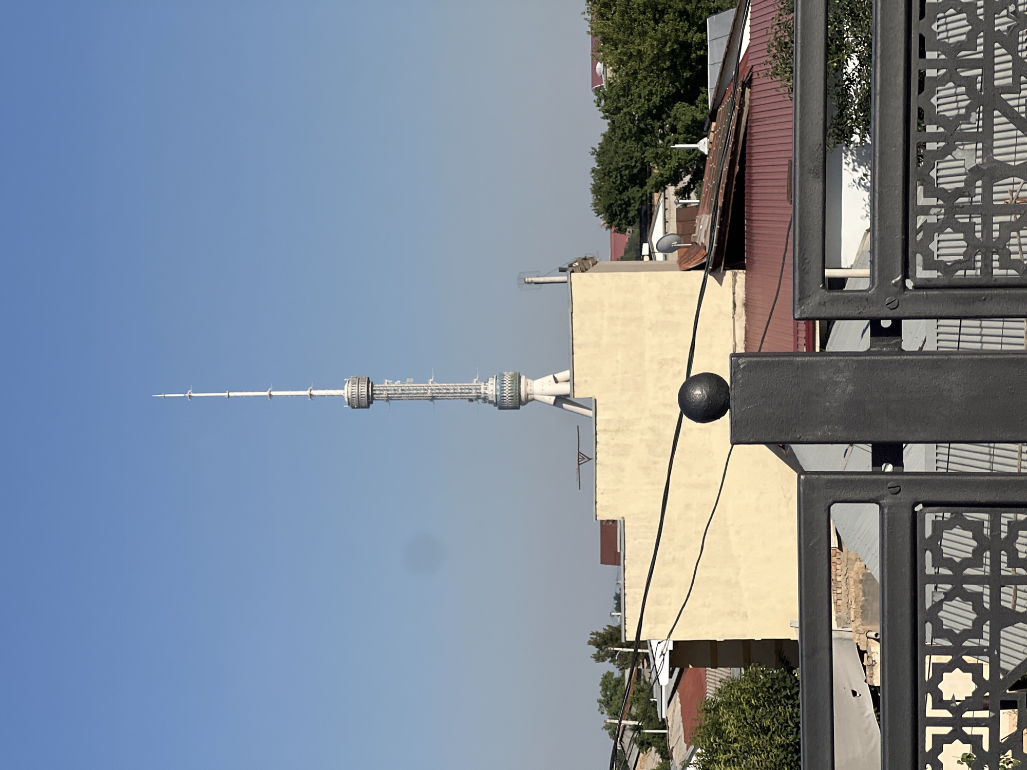 The image captures a view of the Tashkent Television Tower in Tashkent, Uzbekistan, on a clear day. The tower, a prominent landmark, stands tall in the background, its white latticework structure reaching towards the blue sky. In the mid-ground, a light-colored building with a flat roof is visible, with some red-tiled roofs of other buildings peeking out from behind it. Trees with lush green foliage dot the landscape. The foreground features a black metal fence with an intricate geometric pattern, and a black spherical ornament on a post. The scene depicts a sunny urban environment with a mix of modern and traditional architecture.