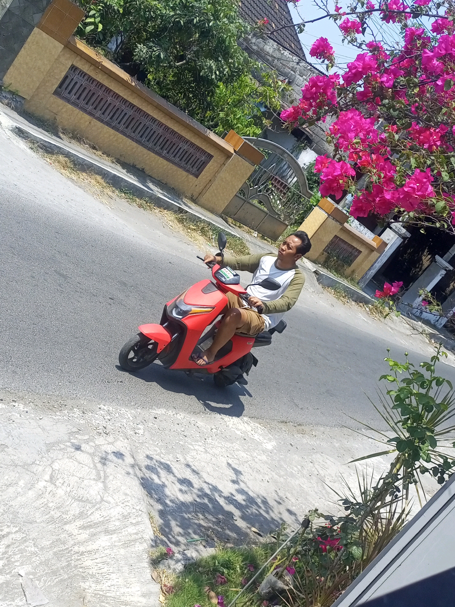 In a sunlit outdoor scene, a young man with dark hair is riding a red electric scooter on a paved road in Ngasem, Indonesia. He is wearing a long-sleeved white and green shirt and khaki shorts, with his right hand on the scooter's handlebars. The scooter has a black mirror and a green sticker on the front. The road is bordered by a concrete sidewalk and a yellow wall with decorative windows and an iron gate. A tree with vibrant pink bougainvillea flowers is visible in the upper right portion of the image, casting shadows on the road. Green foliage and a tall plant with spiky leaves are also present on the right side. The background features a building with a dark roof and stone walls.