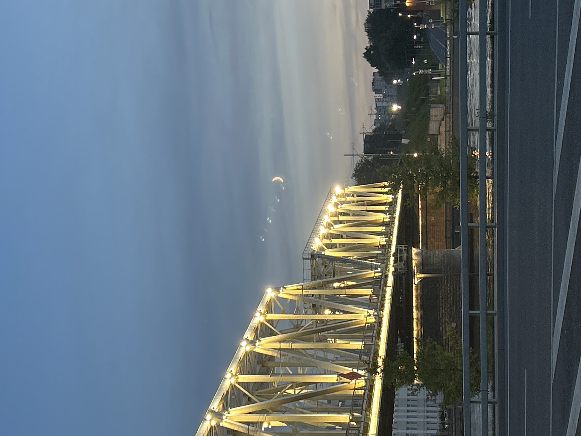 The image shows a yellow metal truss bridge illuminated by lights at dusk or dawn, spanning a body of water. The sky is a gradient of blue and grey, with a crescent moon visible in the upper right portion of the frame. Buildings and trees line the far bank of the river, and streetlights are visible in the distance. The foreground consists of a road with painted white lines and a metal railing separating it from the river. There are no people or animals in the image.