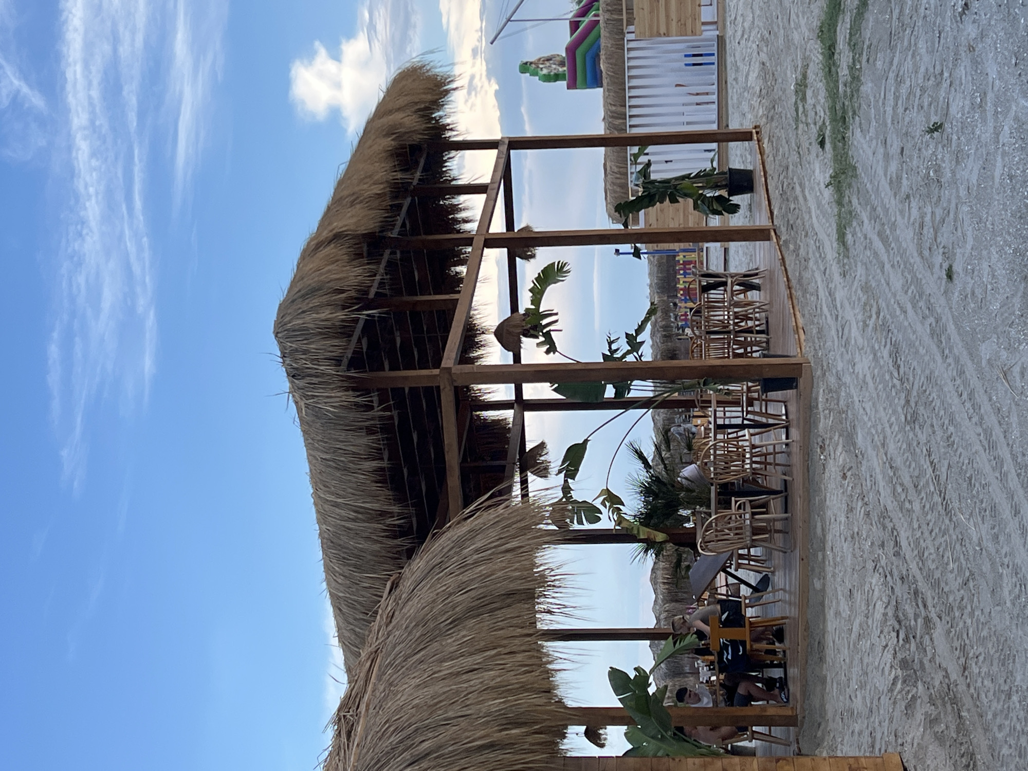 A beachside restaurant with thatched-roof pavilions is depicted under a cloudy sky in Năvodari, Romania. The scene features wooden tables and chairs set on a sandy ground, with several patrons seated and dining. Tropical plants are arranged around the seating area, adding to the ambiance. In the background, an inflatable playground structure and a white wooden fence are visible, suggesting a family-friendly environment. The overall impression is one of a relaxed, outdoor dining experience on a coastal resort.