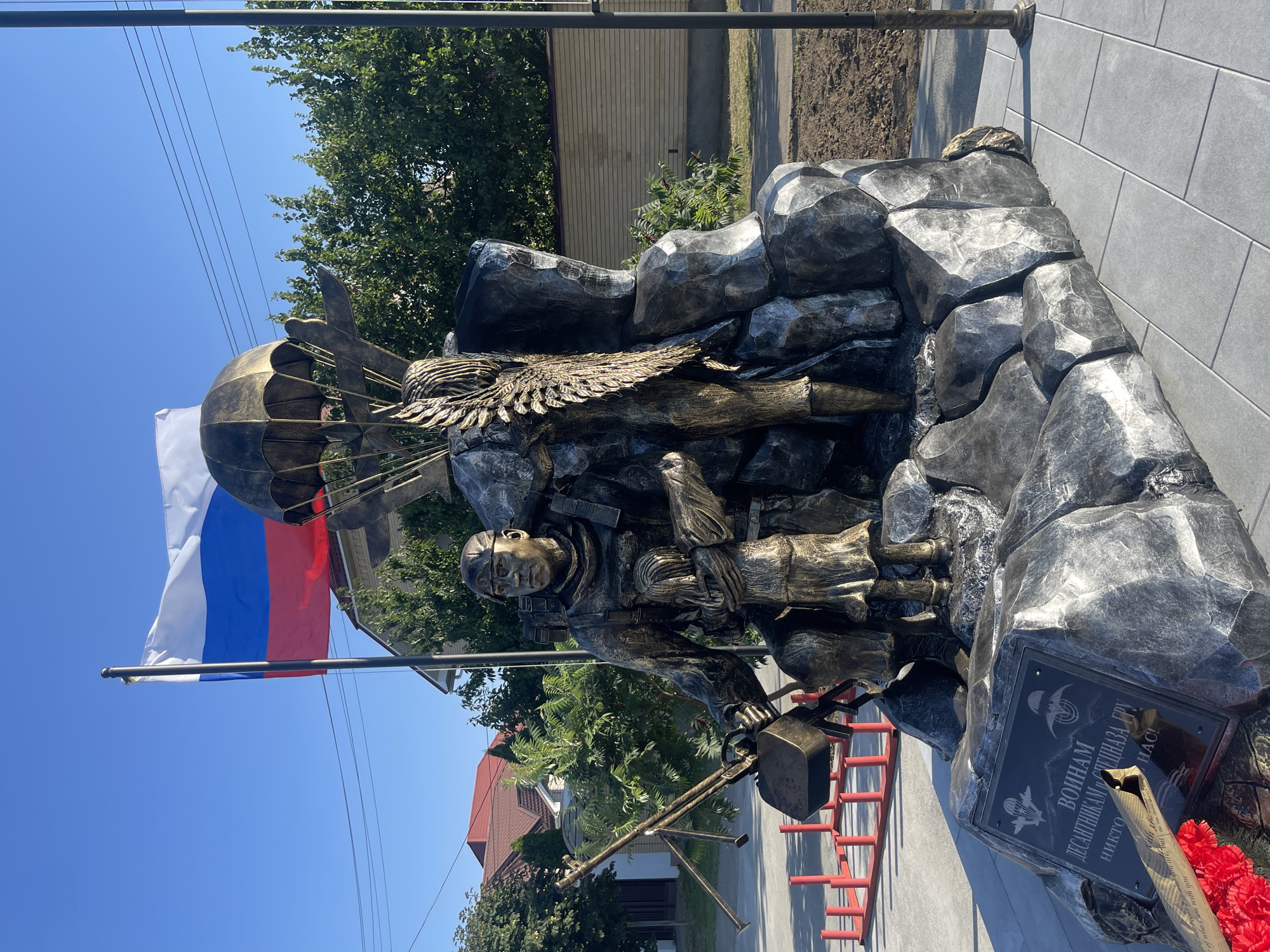This media file showcases a memorial sculpture dedicated to paratroopers, located in Budyonnovsk, Russia. The sculpture depicts a paratrooper in mid-descent, with a parachute canopy partially visible. The figure is rendered in a metallic bronze finish, with intricate details in its uniform and equipment. To the left of the paratrooper, a Russian flag is prominently displayed, waving against a clear blue sky. The sculpture is set against a backdrop of rocky formations, also in a metallic finish, suggesting a rugged terrain.
A small plaque is affixed to the base of the sculpture, bearing an inscription. While not fully legible in the provided images, some text is visible, including the words "ВОЙНАМ" (To the soldiers) and what appears to be "ДЕСАНТНИКАМ" (to the paratroopers). The base of the memorial is adorned with a wreath of red artificial flowers. In the background, trees and buildings are visible, suggesting an outdoor urban or semi-urban setting. The lighting indicates it is daytime, likely late morning or early afternoon, with bright sunlight casting shadows. The overall mood conveyed is solemn and commemorative, honoring the bravery and service of paratroopers.