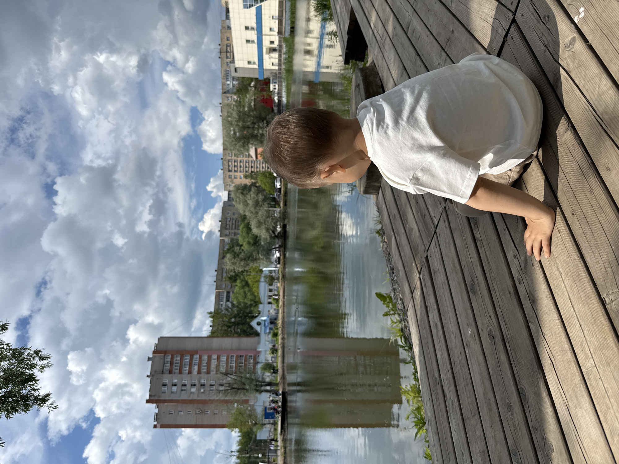 A child in a white t-shirt and light-colored trousers is positioned on a wooden deck, leaning forward and looking intently into a body of water. The child's left hand is resting on the wooden planks near the edge of the deck.
The scene depicts an outdoor environment, likely a park or promenade beside a pond or river, in Rusanovo, Russia. The wooden deck, made of light brown planks, occupies the foreground and right side of the image. To the left of the deck lies the calm water, which beautifully reflects the bright, cloudy sky and the distant urban landscape.
In the background, on the opposite side of the water, several multi-story buildings are visible, ranging from what appear to be apartment blocks to possibly commercial structures. Lush green trees and vegetation line the banks. The sky is a prominent feature, filled with large, fluffy white cumulus clouds set against patches of blue. The time of day appears to be sunny midday or early afternoon, with strong sunlight casting distinct shadows on the wooden deck. There is no visible text in the image. The child's posture conveys a sense of curiosity as they observe something in the water.