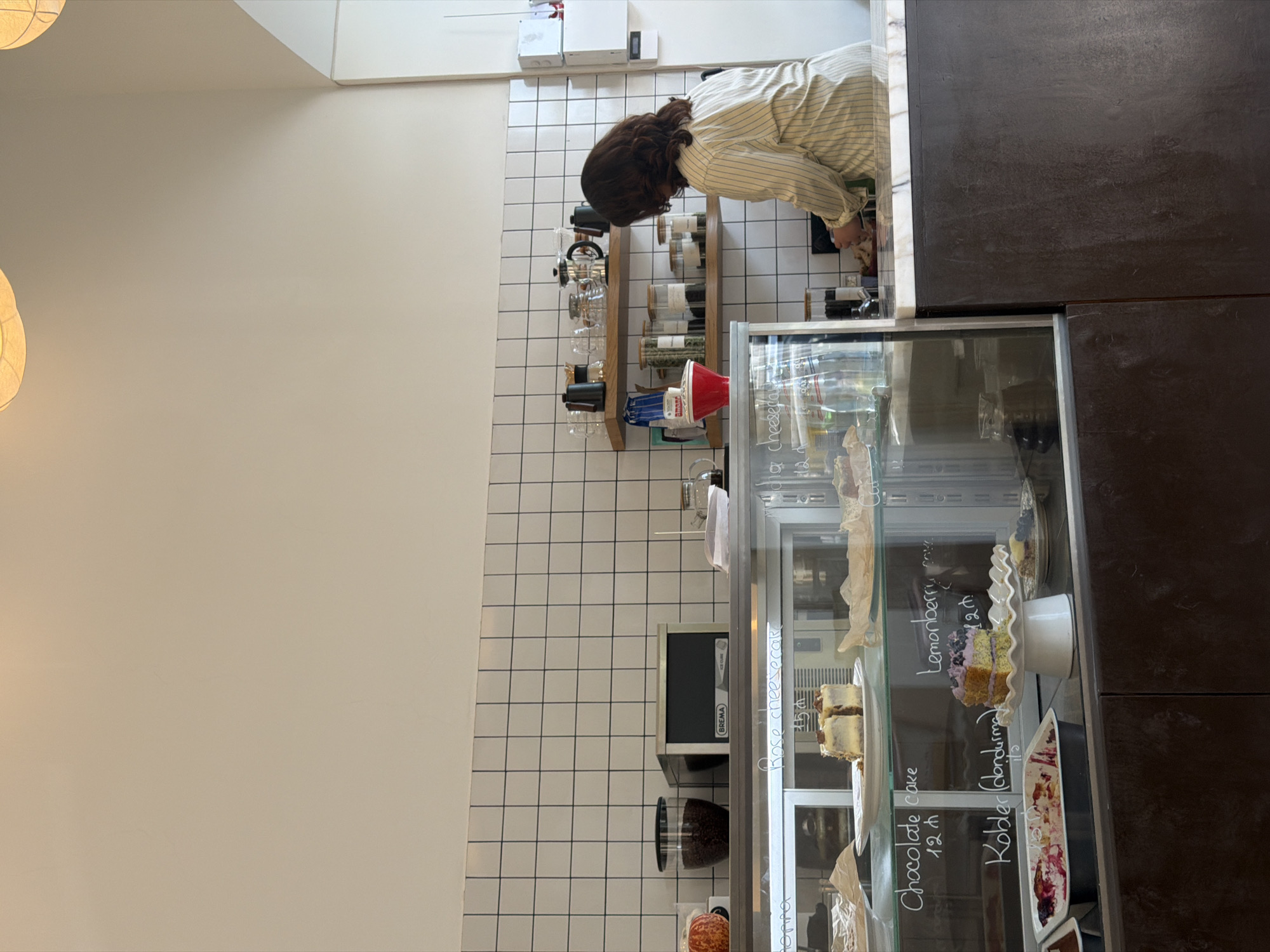 The image shows the interior of a cafe. A woman wearing a striped shirt is working behind the counter, preparing food. In the foreground, there is a display case filled with various desserts, including cakes and pastries. The cafe has a modern, tiled wall and shelves stocked with coffee-making equipment and jars of ingredients. The overall atmosphere appears clean and organized, suggesting a pleasant customer experience.