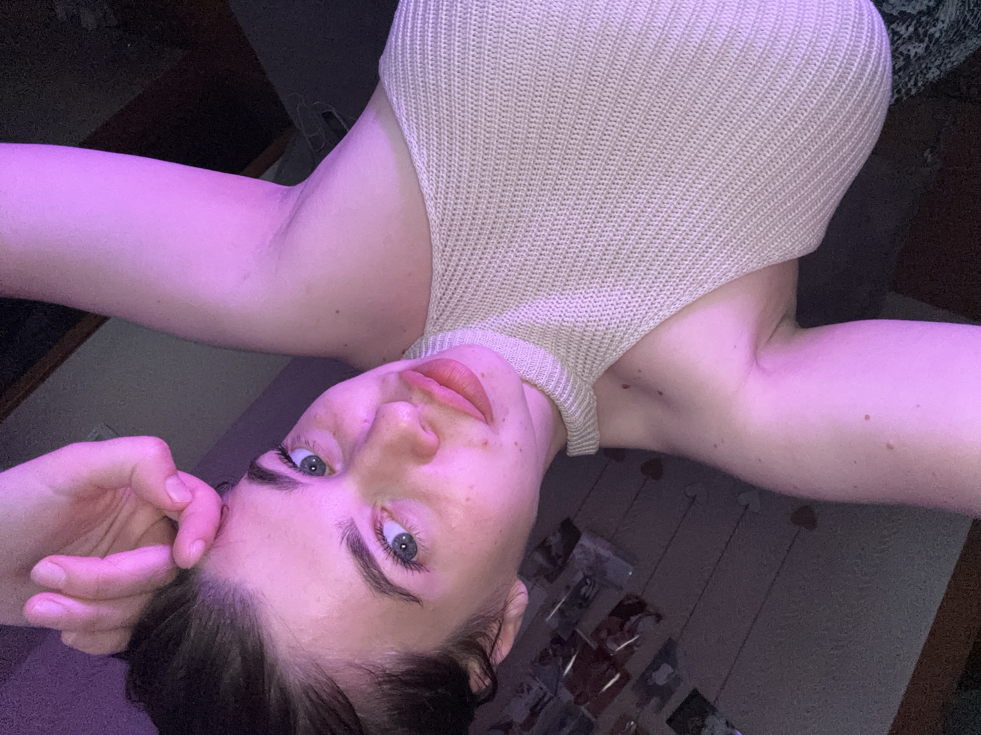 The image shows a young woman with light skin, blue eyes, and dark hair. She is wearing a light-colored, sleeveless knit top. Her left hand is touching her forehead, and her right arm is visible. The lighting in the image is predominantly pink, suggesting a colored light source. The background appears to be an indoor setting with a wooden floor and some decorative elements on the wall. There is no visible indication of the city of Мещёры, Russia, in the image.