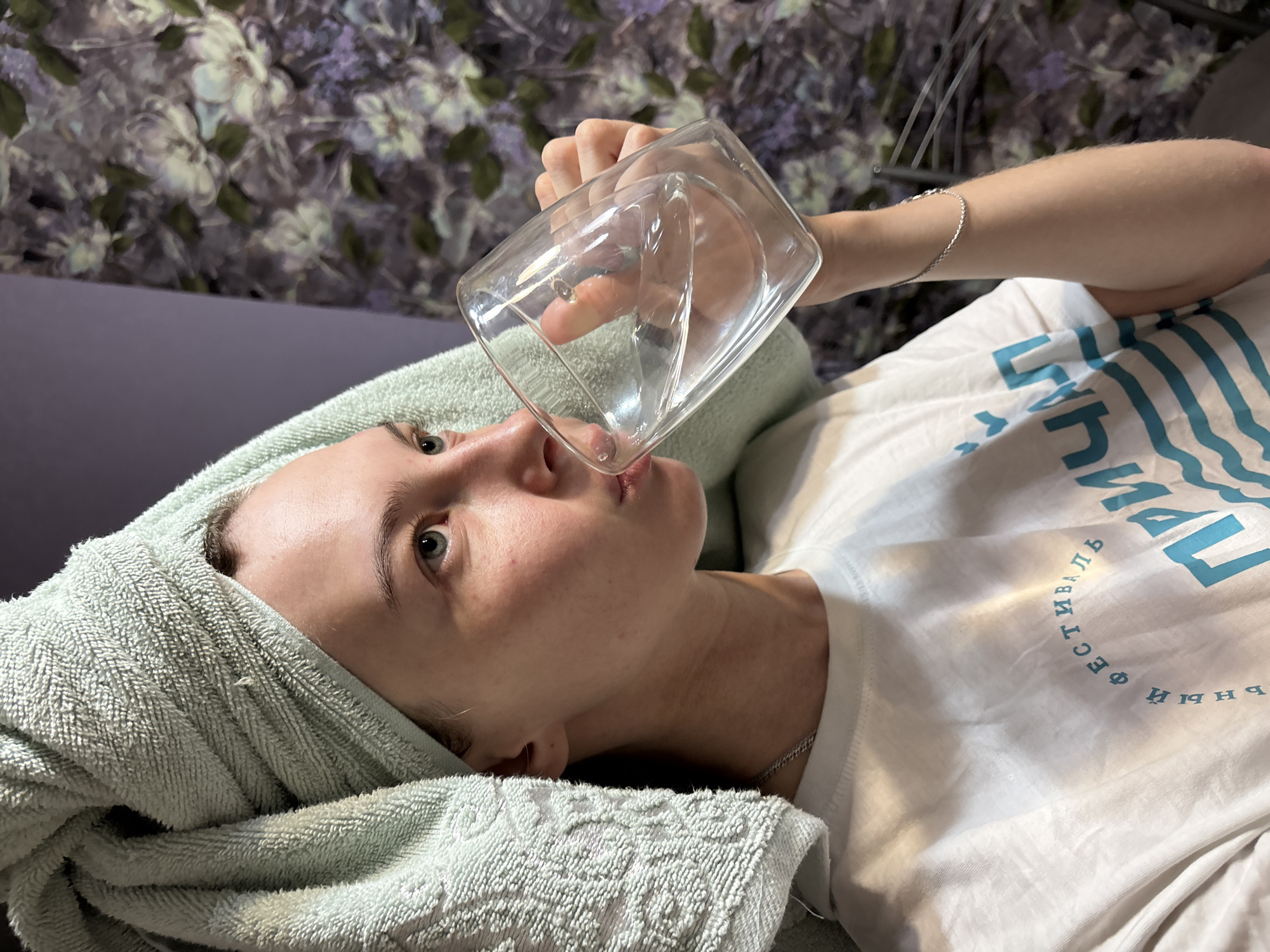 In this close-up, vertical, over-the-shoulder shot, a young woman with light skin and blue eyes drinks from a clear, double-walled glass of water. Her hair is wrapped in a light blue towel, and she wears a white t-shirt with blue text that appears to be in Cyrillic script. The background is a floral wallpaper with muted colors. The scene does not provide any discernible information about a specific location within the city of Perm, Russia.