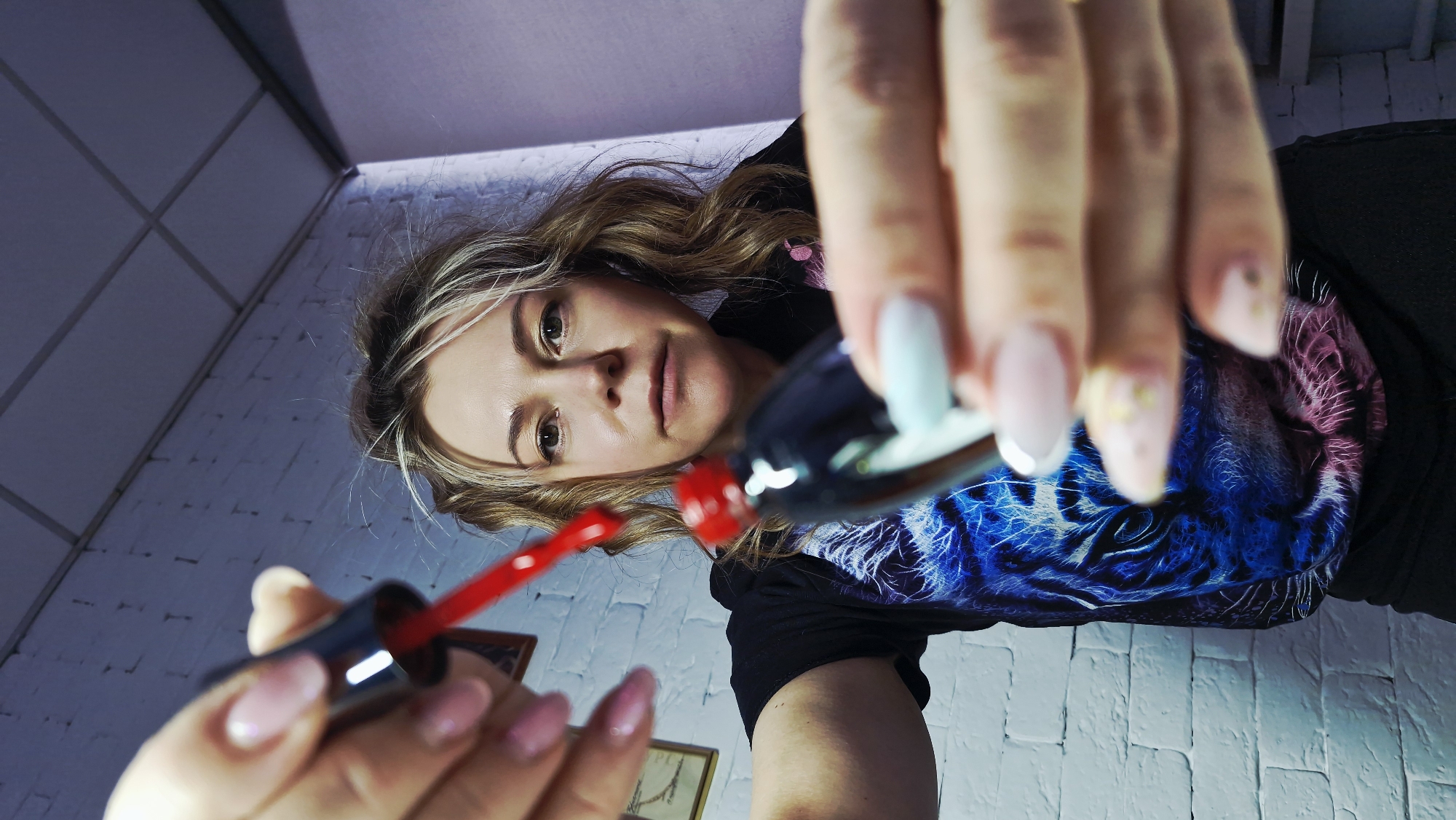 This low-angle, close-up shot captures a woman looking directly into the camera from a downward perspective. Her face, positioned in the upper center, is in focus, revealing light-colored hair with highlights and a neutral, composed expression. She is wearing a dark t-shirt adorned with a striking, colorful graphic of a tiger's head, rendered in shades of blue and pink/purple.
In the foreground, her manicured hands are prominently displayed, holding a bottle of vibrant red nail polish with the brush extended. Her nails feature various designs, including light pink with white tips and some adorned with glitter or star-like embellishments. The hands and nail polish are slightly out of focus due to their proximity to the camera, creating a shallow depth of field that emphasizes the woman's face.
The activity depicted is the preparation for or demonstration of nail polish application. The setting appears to be an indoor room, indicated by a light-colored, possibly white or light grey, brick-patterned wall in the background and a grid-patterned ceiling above. The lighting seems to be artificial. There are no specific visual cues in the image to directly confirm the provided location context of Otchor, Russia, beyond it being an interior space. No text is visible in the image. The time of day is not discernible due to the indoor artificial lighting.