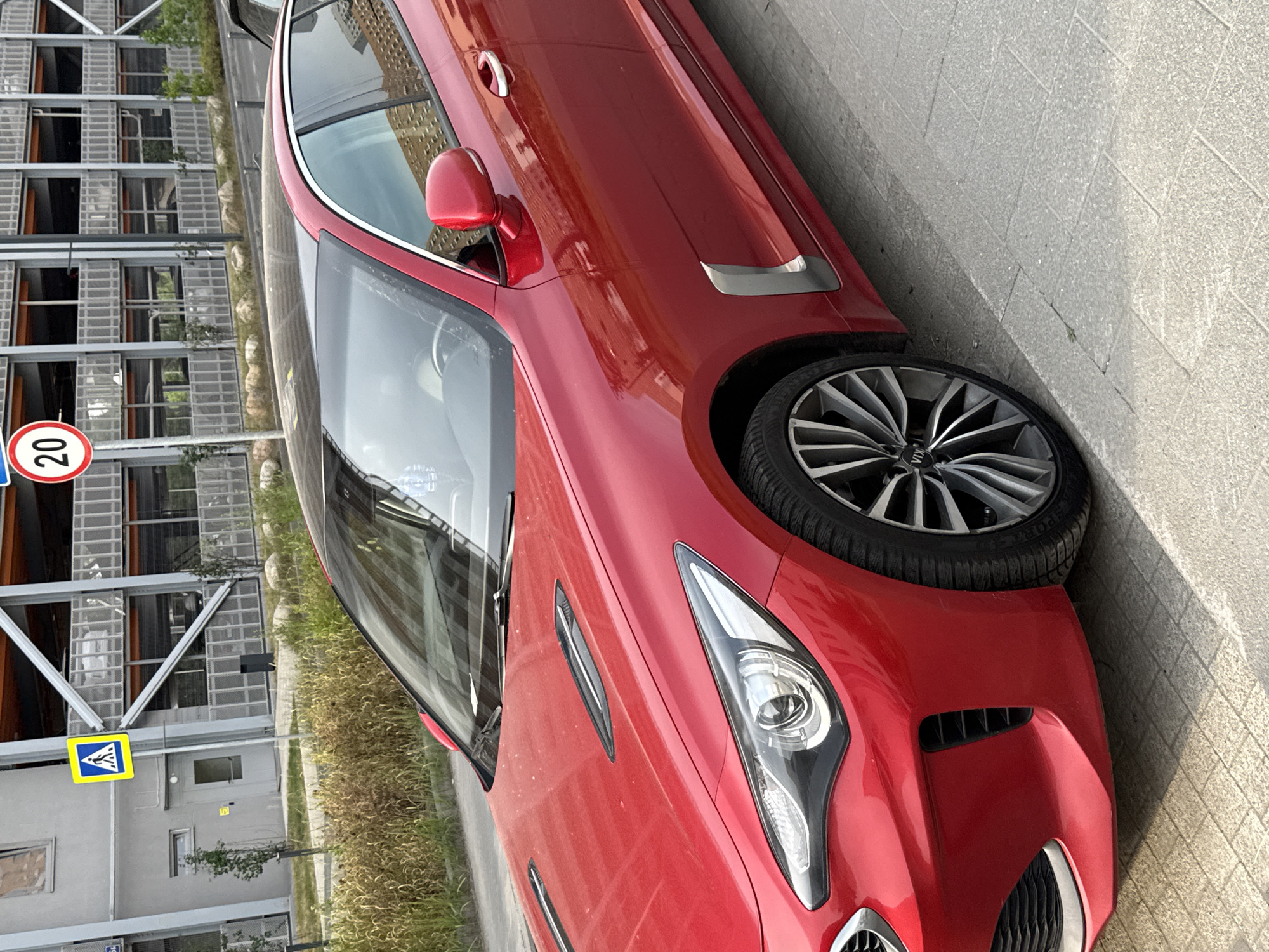 A red Kia Stinger is parked on a gray tiled sidewalk next to a multi-story parking garage with gray metal grating. A yellow pedestrian crossing sign is visible on the left side of the image, as is a circular speed limit sign showing "20". Dry, light brown grass and small rocks line the edge of the sidewalk. The car's hood has two dark vent-like accents. The wheels are a silver and black multi-spoke design. The scene is outdoors during the daytime.