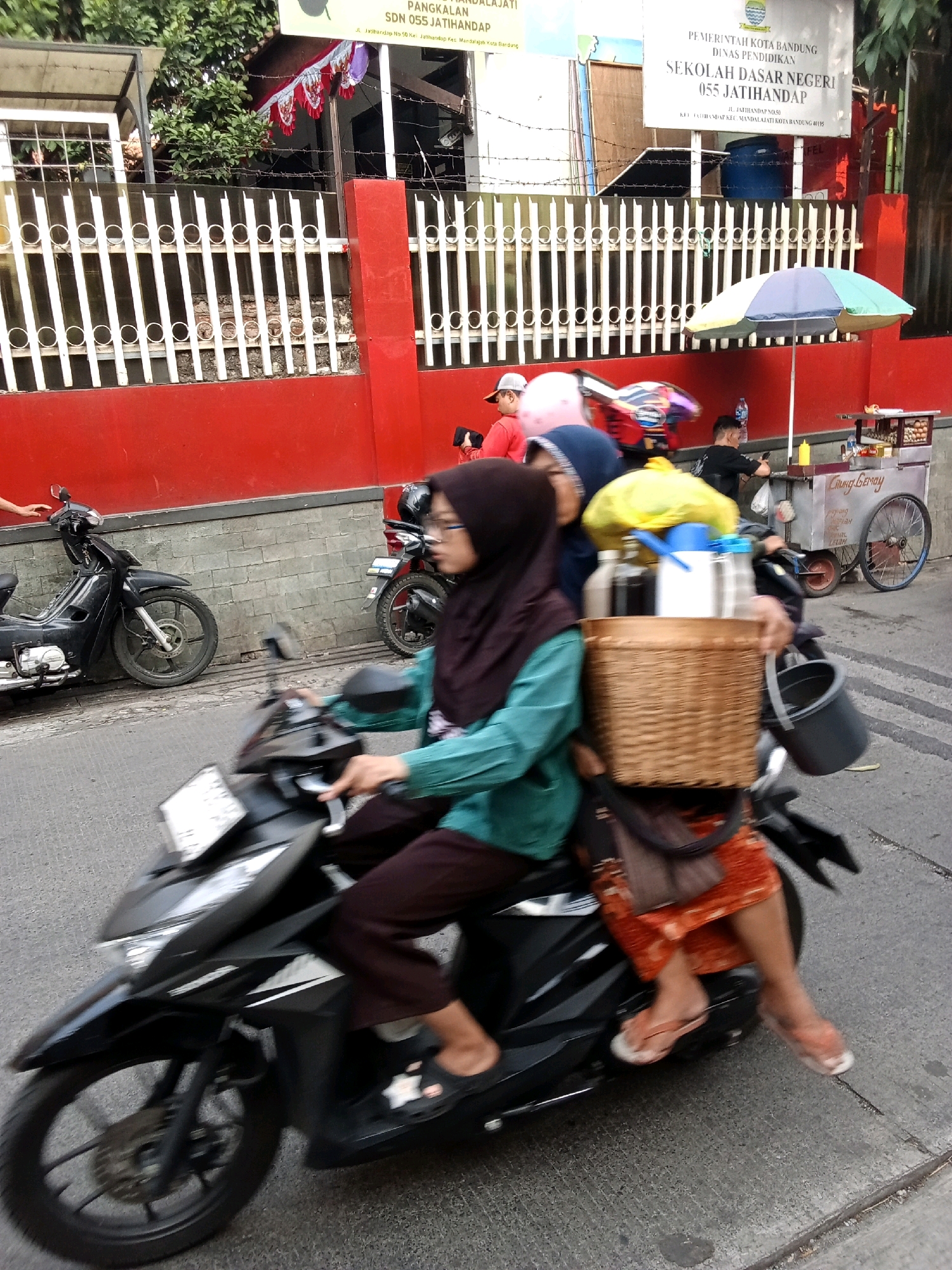 The image depicts a street scene in Bandung City, Indonesia. In the foreground, a woman wearing a hijab and glasses is riding a black motorcycle. She is dressed in a teal shirt and dark brown pants. On the back of the motorcycle, a basket is attached, containing several bottles and a gray bucket. The woman is carrying a brown handbag over her shoulder. Another person, wearing a blue hijab, is seated behind her. The motorcycle is in motion, indicated by the blur of the wheels and the surroundings.
In the background, a red wall runs along the street. A white fence with vertical bars is visible in front of the wall. To the right of the fence, a sign reads "PEMERINTAH KOTA BANDUNG DINAS PENDIDIKAN SEKOLAH DASAR NEGERI 055 JATIHANDAP." This indicates the location is a primary school named SDN 055 Jatihandap, under the Bandung City Government's Education Department. Further in the background, a food cart with a wheel is visible, with an umbrella providing shade. There are also other people and motorcycles in the distance, suggesting a bustling street environment. Trees are visible above the fence on the left side of the image.