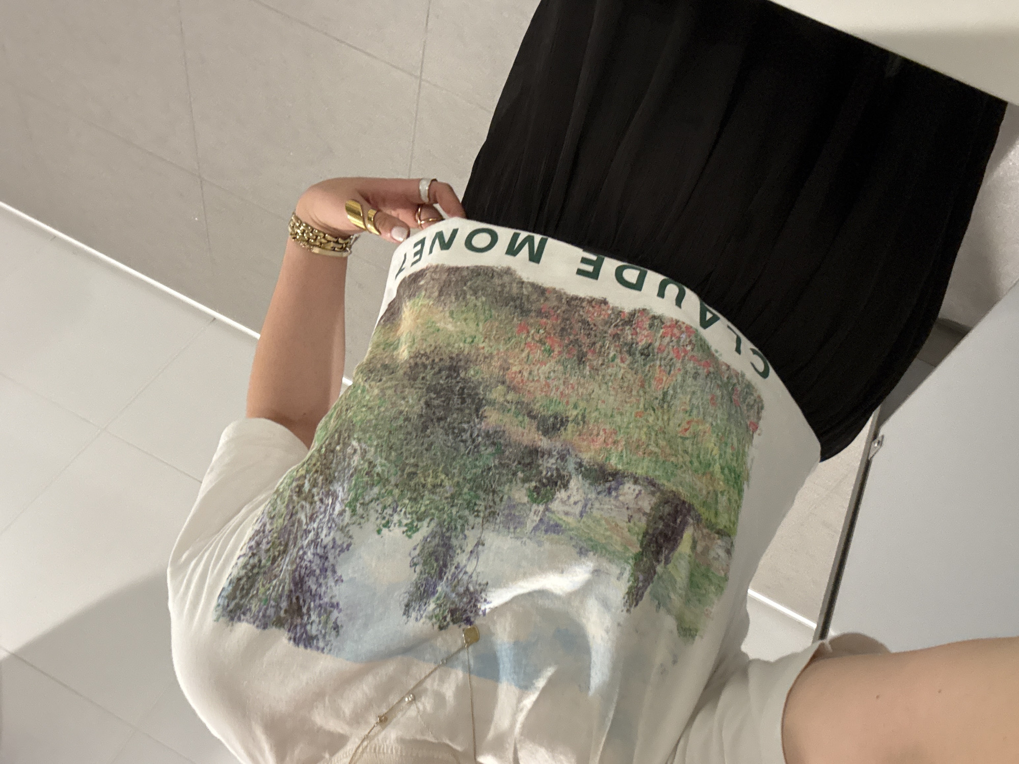 The image depicts a person wearing a white t-shirt with a print of a Claude Monet painting and the artist's name in bold, green letters. They are also wearing a black skirt. The person has gold jewelry on their hand and wrist. The background appears to be a tiled floor and a white wall. The photograph may have been taken in a restroom. The location (Mexico City, Mexico) cannot be determined from the image.