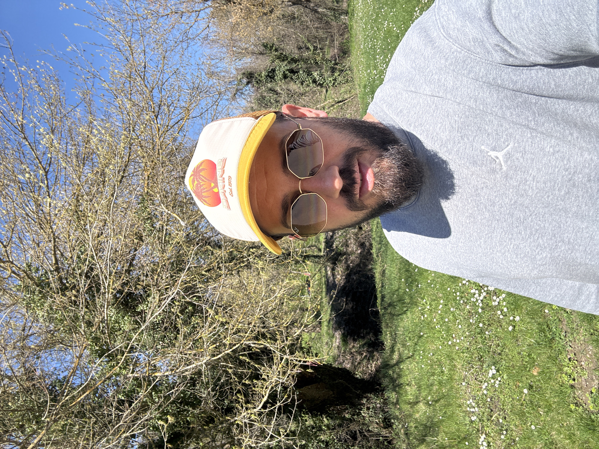 A man is pictured outdoors, likely in a park or green space. He wears a light grey sweatshirt with a small white logo on the left chest area, a white and yellow baseball cap with a Dakine logo, and brown-tinted sunglasses. The background features trees with bare branches, some with budding leaves, indicating early spring. Green grass speckled with small white flowers is visible. The location is possibly in the city of Pleven, Bulgaria, but without further details, this cannot be confirmed. The angle of the photo suggests it may be a selfie.