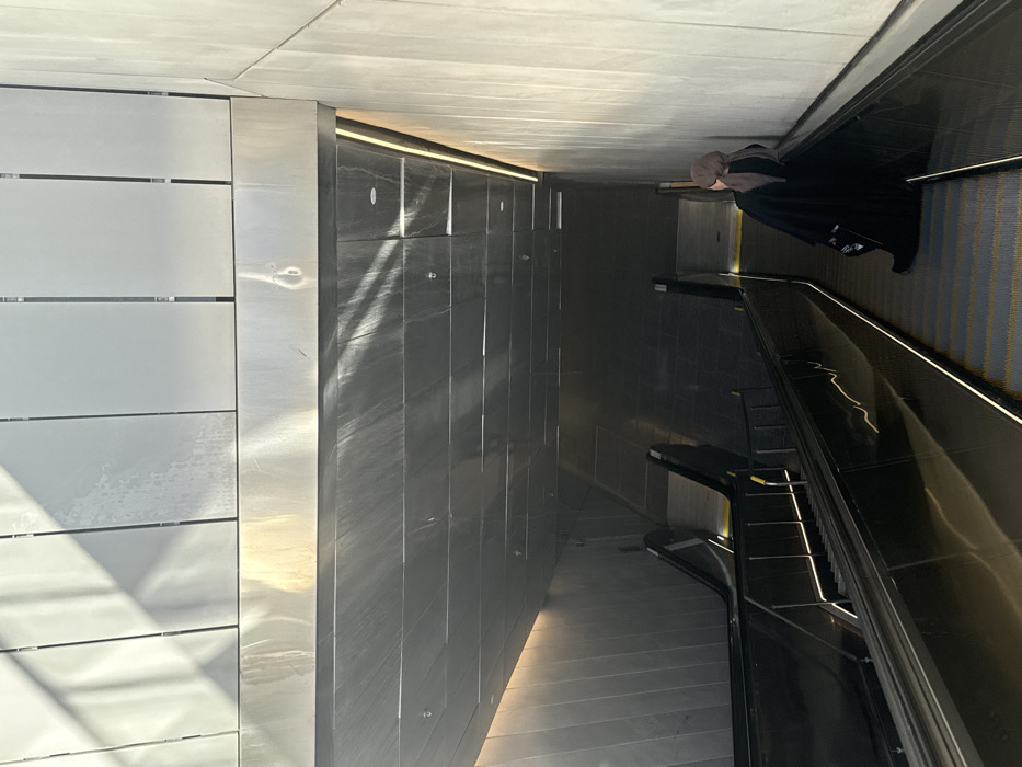 An indoor scene shows a person riding an escalator in a modern building. The walls are lined with rectangular panels in light and dark metallic hues. The escalator has a black handrail and steps with a visible yellow striped pattern. The overall aesthetic is clean and minimalist, with light playing off the metallic surfaces.