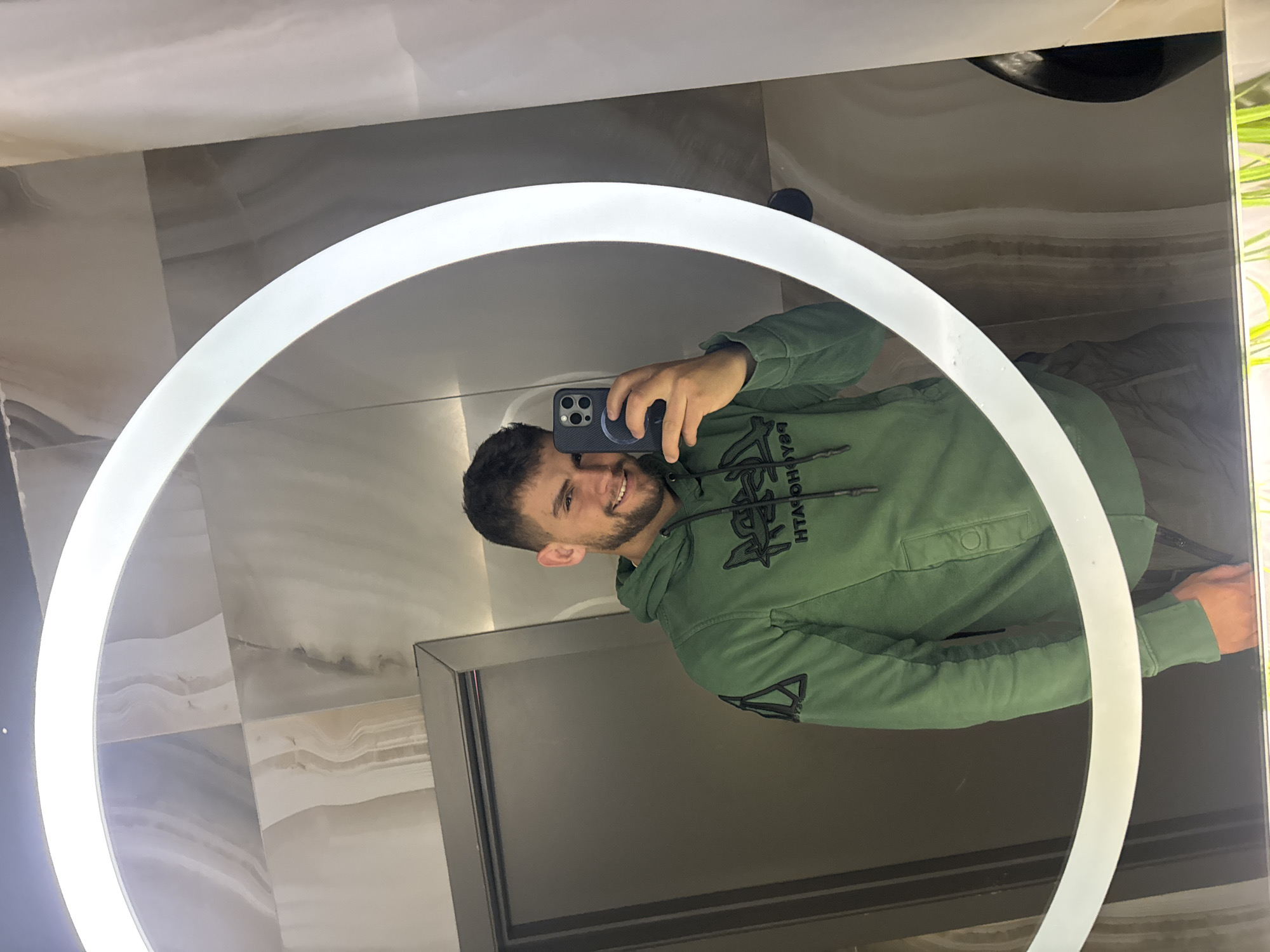 This image is a mirror selfie captured by a young man indoors, set in Sofia, Bulgaria. The man, who has dark hair and a beard, is depicted from the chest up, smiling directly at the camera while holding a dark-cased smartphone. He is wearing a green hooded sweatshirt featuring an inverted, stylized text logo across the chest, which, when un-inverted, reads "STÜSSY." A smaller, inverted triangular graphic is also visible on his left sleeve. The selfie is taken in a circular mirror framed by a bright, illuminated LED light ring. The background consists of modern, light-toned wall tiles with a subtle veined pattern, possibly marble or stone. A dark, framed section, appearing to be part of furniture or a door, is reflected in the lower portion of the mirror. A hint of green, suggesting foliage or a decorative plant, can be seen on the right edge of the frame. The man's joyful expression is evident from his smile.