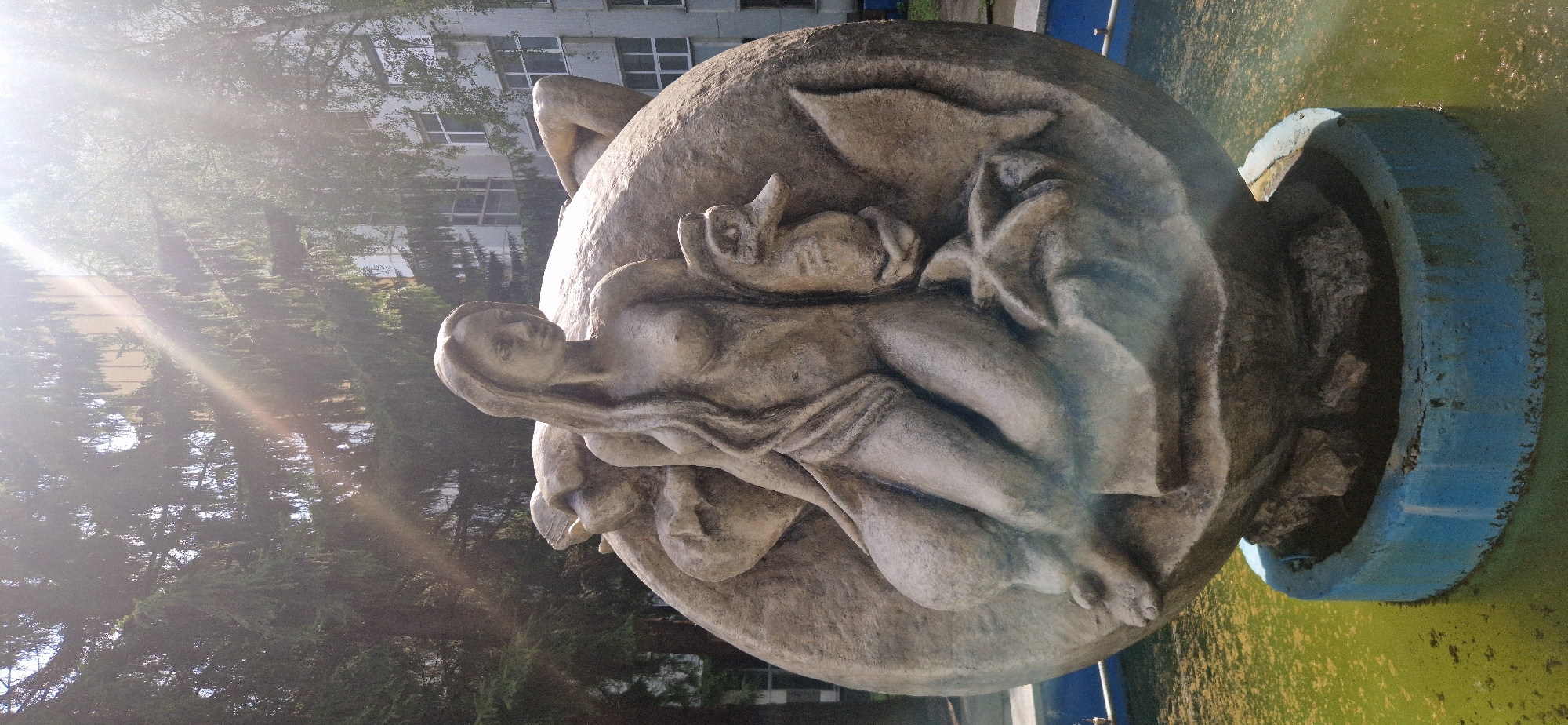 The image shows a fountain sculpture located in Krasnoobsk, Russia. The sculpture is made of stone and depicts a nude female figure sitting on a large, round object that resembles a globe. She has long, wavy hair and is draped with a cloth. Beside her, on the round object, are carvings of a seahorse, a starfish, and what appears to be a fish tail. The sculpture is partially submerged in a pool of green water, and the base of the fountain is a blue concrete ring. In the background, there are tall trees with dense foliage and a building with windows. Sunlight is casting rays through the trees, creating lens flare effects in the image.