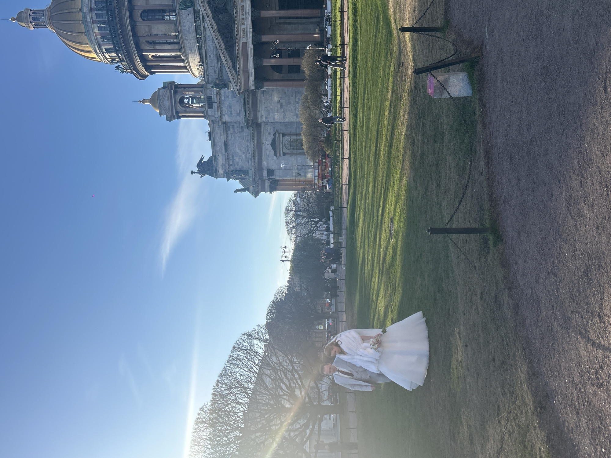 In the foreground, a bride and groom pose on the grass. The bride is wearing a white wedding dress and veil. The groom is wearing a light gray suit with a tie. They are standing near a row of trimmed trees with a bright sun behind them.
In the background, St. Isaac's Cathedral dominates the landscape, a notable landmark in Saint Petersburg, Russia. People can be seen walking and congregating near the cathedral. The sky is clear and blue. A section of a gravel path is visible in the lower right corner. A set of chained posts lines the edge of the lawn.