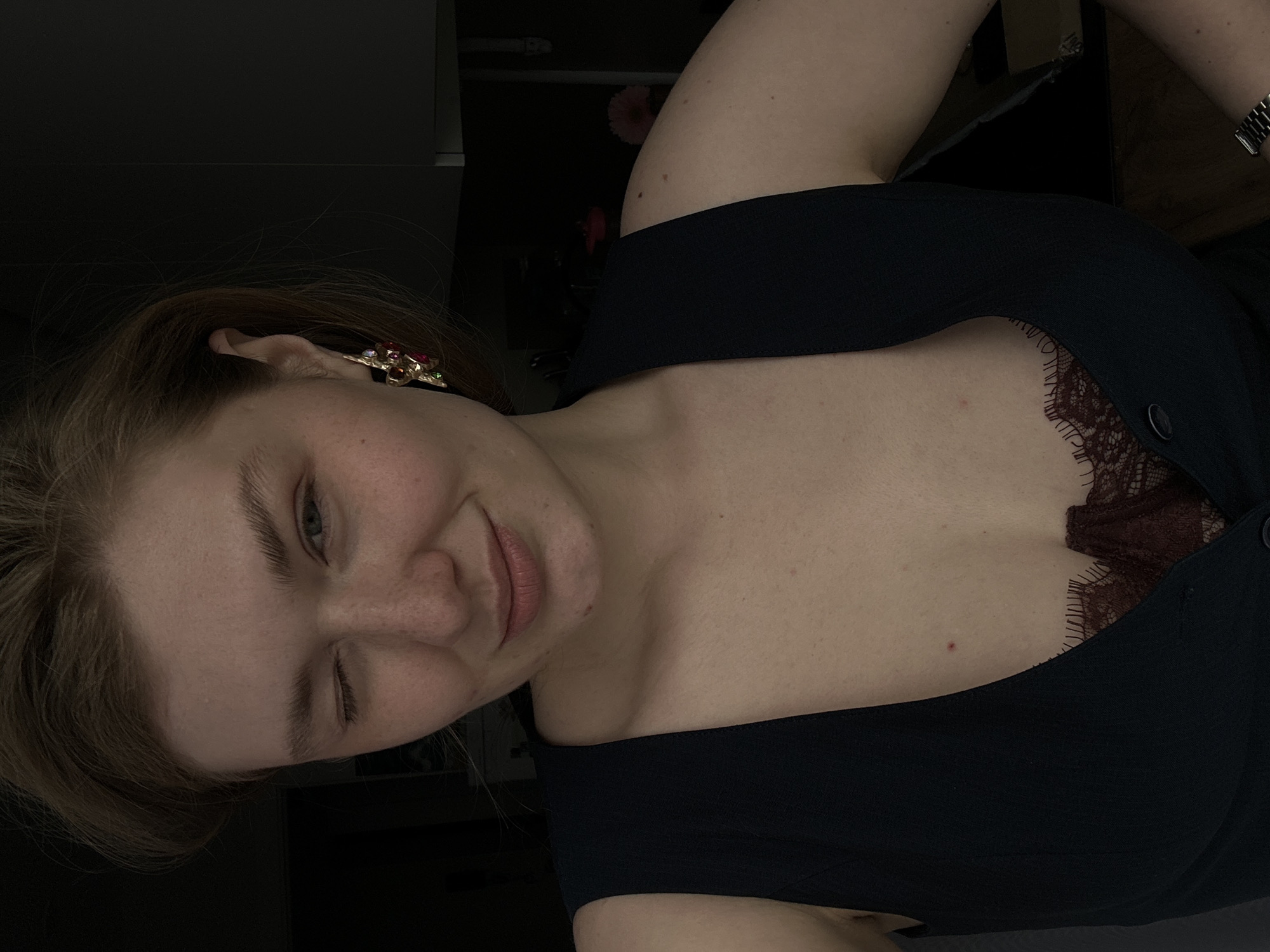 A fair-skinned woman winks at the camera. She is wearing a black garment, underneath of which is a deep red top with lace detail, and an ornate gold earring. The setting is a darkened interior with indistinct objects in the background. There is no indication this image was taken in Kazan, Russia.