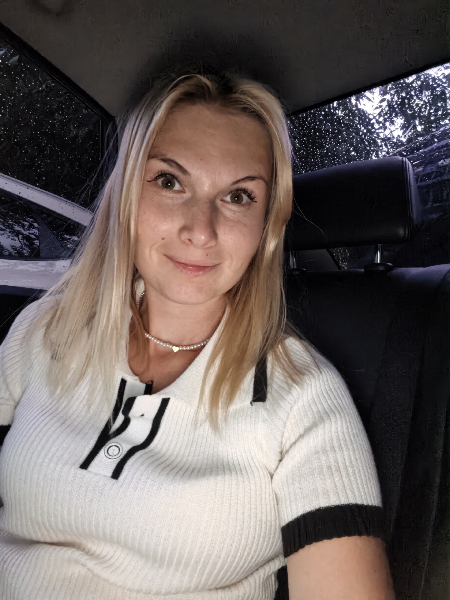 A woman with blonde hair and light skin is captured in a close-up, front-facing shot. She is seated inside a vehicle, indicated by the leather headrest of a seat behind her and a window with visible raindrops on the exterior, suggesting it is raining outside. The woman is wearing a white, ribbed sweater with black trim and a small pearl necklace with a gold heart pendant. Her makeup includes winged eyeliner. The background appears dark and slightly blurred, consistent with the interior of a car at night or in low light conditions. The image does not provide any specific details that would link it to Sterlitamak, Russia, or any other city.