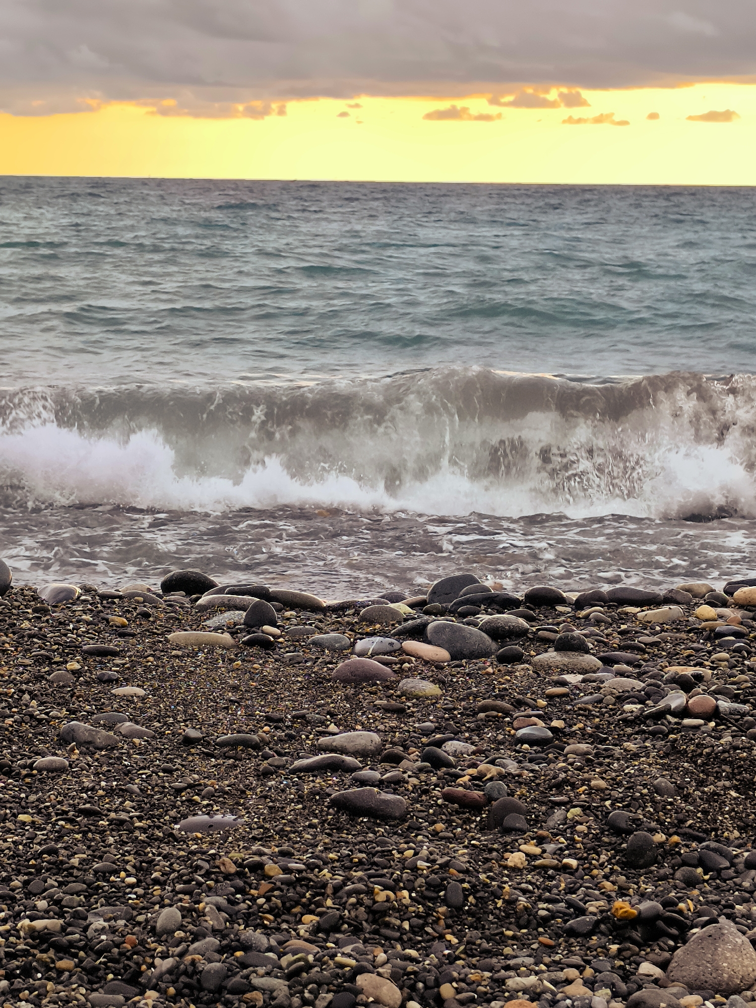 The image depicts a rocky beach with the sea and a cloudy sky. The foreground is filled with an abundance of dark, smooth pebbles of various sizes, interspersed with smaller stones and some lighter-colored, smooth stones. A wave is breaking in the middle ground, with white foam crashing onto the shore. The sea is a choppy, dark blue-gray color. The horizon line separates the sea from the sky, which is dominated by a band of bright yellow and orange, suggesting either sunrise or sunset, with dark, heavy clouds above. The scene is devoid of any people, structures, or vegetation. The location is identified as Zemo Kvariati, Georgia, suggesting a coastal setting in that region.
