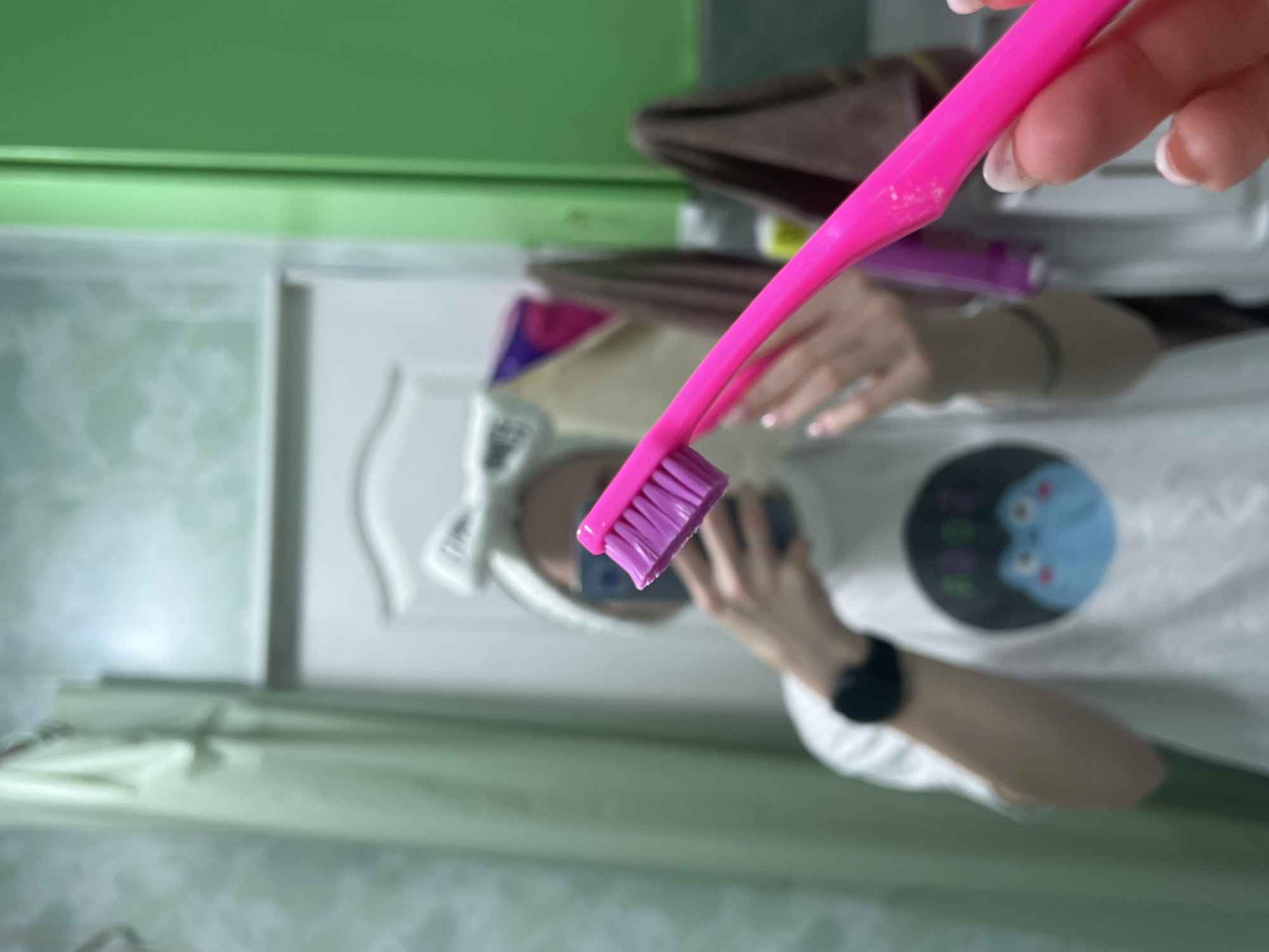 A person is taking a selfie in a bathroom mirror. The person is wearing a white shirt with a blue cartoon design. A pink toothbrush is held up in front of the mirror. The bathroom has green walls and a white cabinet. A towel is visible in the background. The location is Chaikovsky, Russia.
