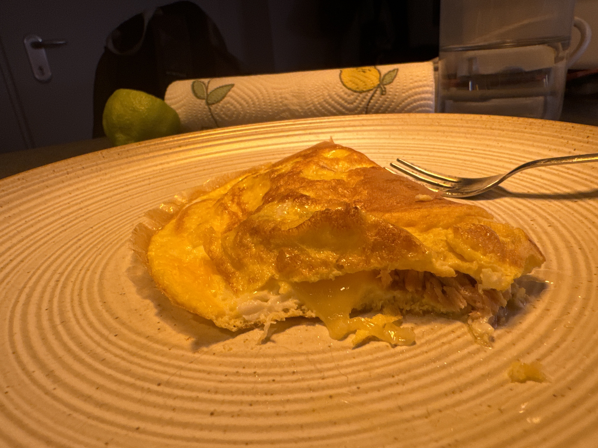 A close-up, low-angle shot shows a partially eaten omelet on a textured, circular plate. The omelet is golden brown and appears to be filled with cheese, which is slightly melted and oozing out. A fork is placed on the right side of the plate, partially resting on the omelet. In the background, out of focus, is a roll of paper towels with a lemon or lime design, a lime, and a glass of water. The setting appears to be a domestic environment, likely a kitchen or dining area, with the warm lighting suggesting it might be nighttime or an indoor setting with artificial light. There is no indication of this scene being in Murcia, Spain.