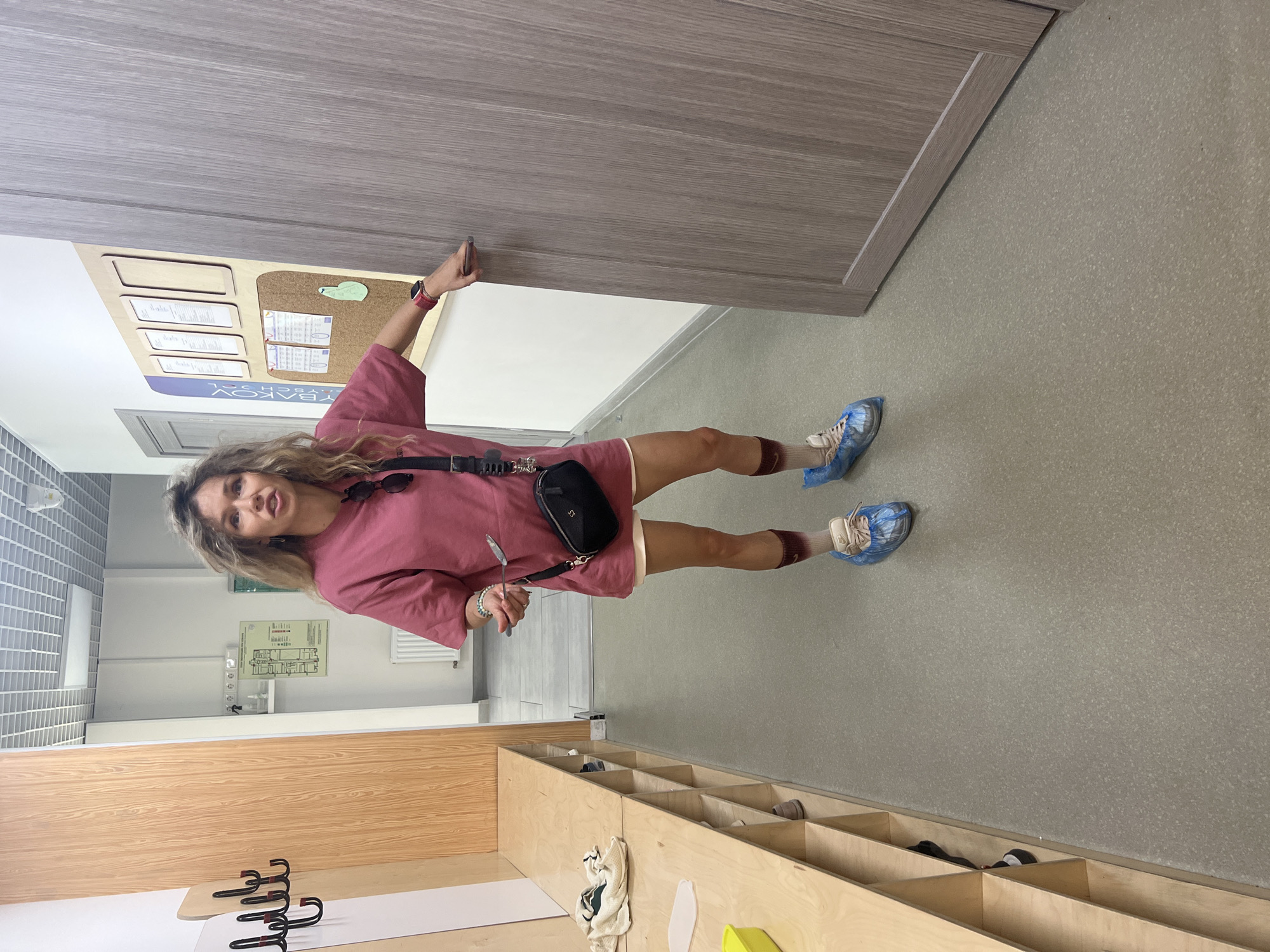 A woman with blonde curly hair stands in a hallway, wearing a pink t-shirt, white shorts, and blue shoe covers. She is holding a metal utensil in her right hand. A black crossbody bag is slung over her left shoulder. To her left is a row of wooden cubbies. To her right is a light brown wooden door that she is reaching for with her right hand. The hallway has a grey tiled floor and white walls. A corkboard with papers is visible on the wall behind her.