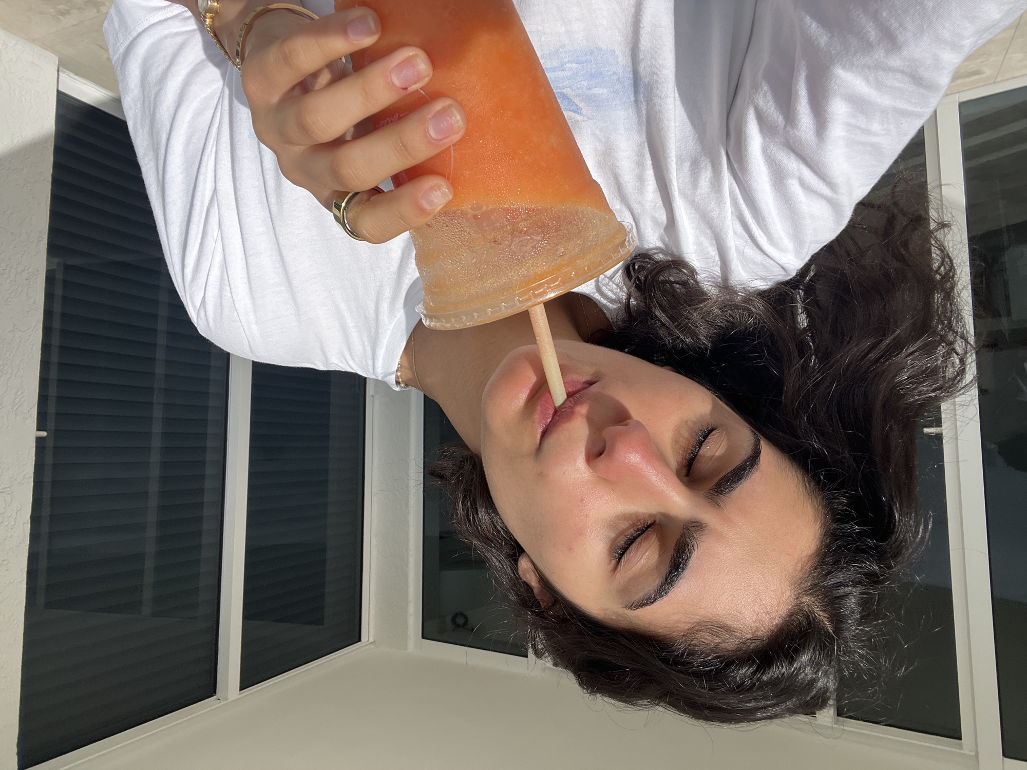 A young adult with dark hair is positioned indoors, holding a large cup of an orange-colored drink with a straw to her lips. She has her eyes closed. She is wearing a white, long-sleeved shirt and a gold bracelet and ring on her left hand. Behind her is a window with dark blinds. The image was taken in Miami Beach, United States of America.