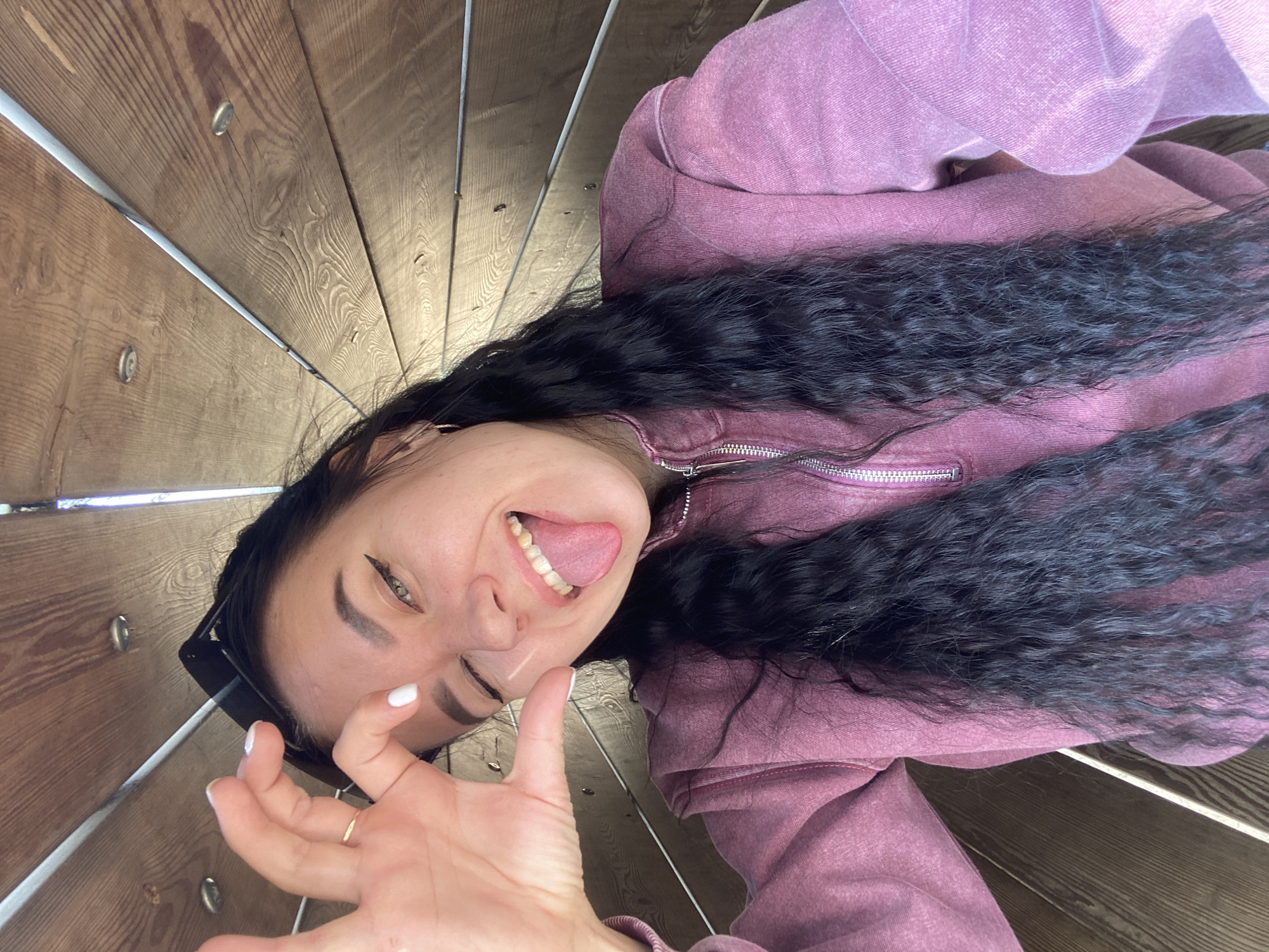 A woman is lying on a wooden surface, looking directly at the camera and sticking out her tongue. She is wearing a maroon zippered hoodie and has long, dark curly hair. Her right hand is raised with her fingers splayed, and she has white nail polish. A ring is visible on her right hand. Sunglasses are pushed back on her head. The background consists of wooden planks arranged in a circular pattern, giving the impression of being inside a large wooden structure. The location is the Rotunda, a distinctive architectural feature in Perm, Russia.