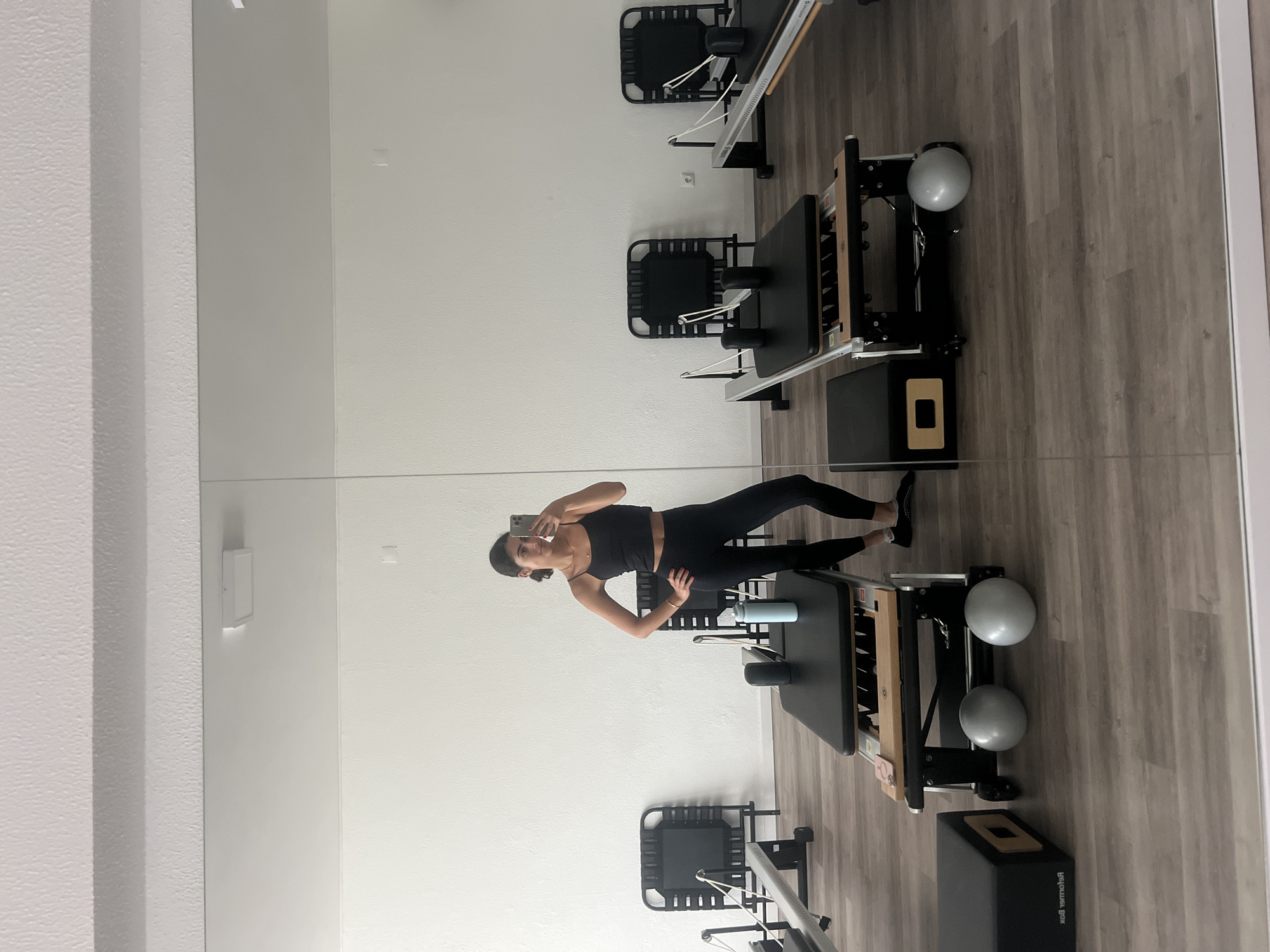 A woman with dark hair tied back is standing in a pilates studio in Matosinhos, Portugal, taking a mirror selfie. She is wearing a black sports bra and black leggings. She is positioned in the center of the frame, with her right hand on her hip and her left hand holding a smartphone.
Behind her, multiple pilates reformers are visible, set up in rows. The studio has white walls and a wooden floor. A large mirror covers a significant portion of the wall behind the woman and the reformers, reflecting the scene. There are several small, light grey pilates balls on the floor near one of the reformers. A light blue water bottle is also on the reformer. To the left of the frame, a black pilates box is visible. The overall lighting is bright, suggesting natural light or well-lit studio conditions. The scene appears clean and organized, indicative of a professional fitness environment.