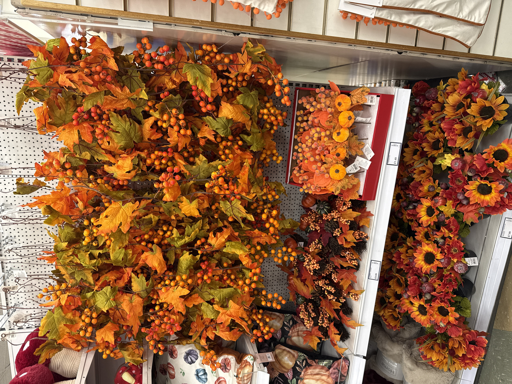 The image depicts a retail display of autumn-themed decorations in a store. The primary focus is on garlands of artificial autumn leaves and berries, predominantly in shades of orange and green, hanging from hooks. Below these garlands, on shelves, are various other decorative items. These include a box filled with small artificial pumpkins and berries, as well as bouquets of artificial sunflowers and other fall flowers in black pots. Several pillows with pumpkin motifs are also visible on a lower shelf, along with a fluffy, light-colored cushion. The environment appears to be a well-lit retail space with shelving units and a textured white wall in the background. There are price tags visible on some of the items, indicating they are for sale. No people are present in the image. The location of this scene is confirmed as Darlington, United Kingdom, based on the visible price tags with GBP currency.