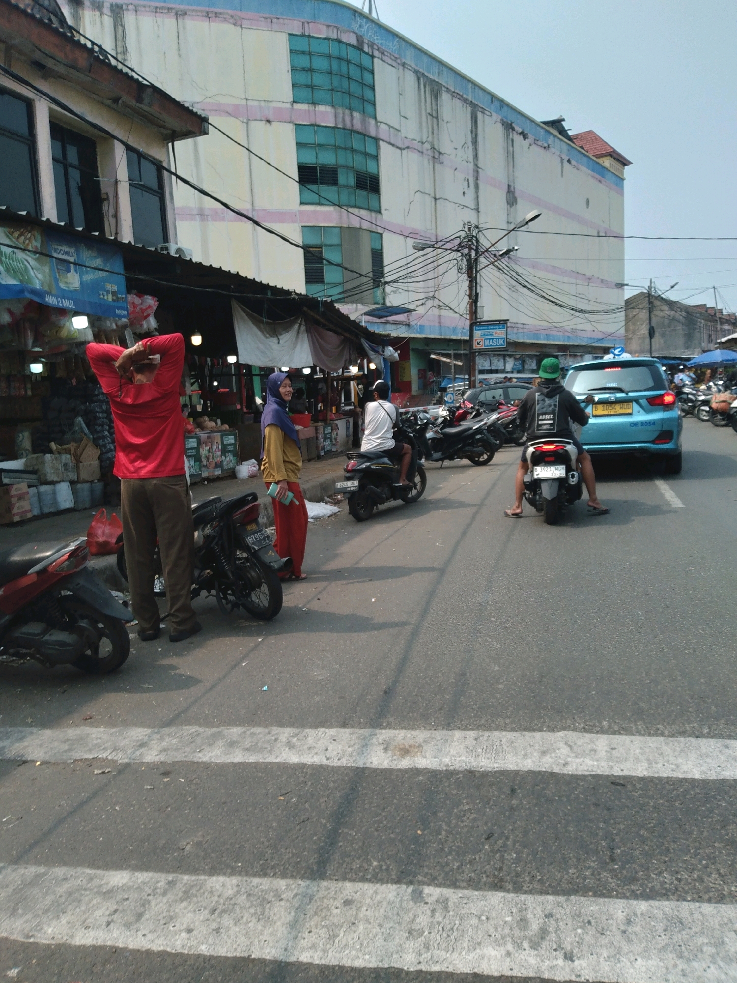 This image captures a street scene in South Tangerang, Indonesia, likely near a market or commercial area, with a large building in the background that has a light-colored facade with pink and blue horizontal stripes. The building appears to be multi-story with multiple windows. In the foreground, a two-lane asphalt road is visible with white pedestrian crossing lines.
Several individuals and vehicles are present. On the left side of the road, a person wearing a red long-sleeved shirt and brown pants stands with their hands behind their head, facing away from the camera. Next to them, a red motorcycle is parked. Further down the street, a woman in a yellow top and red pants, wearing a blue hijab, is standing and holding a blue object.
Several motorcycles are parked and in motion on the road. One black motorcycle is parked in the middle of the scene, and another black motorcycle is seen in motion to the left of the woman in the hijab. In the right half of the image, a blue car with a yellow license plate is on the road. A person wearing a black hoodie and a green hat is riding a black motorcycle behind the blue car. Another person is seen riding a motorcycle on the far right of the image.
The environment suggests a warm, possibly sunny day with the presence of street stalls or shops lining the left side of the road. Overhead power lines are prominent throughout the scene, strung between poles and the buildings. The overall impression is of a busy urban street.