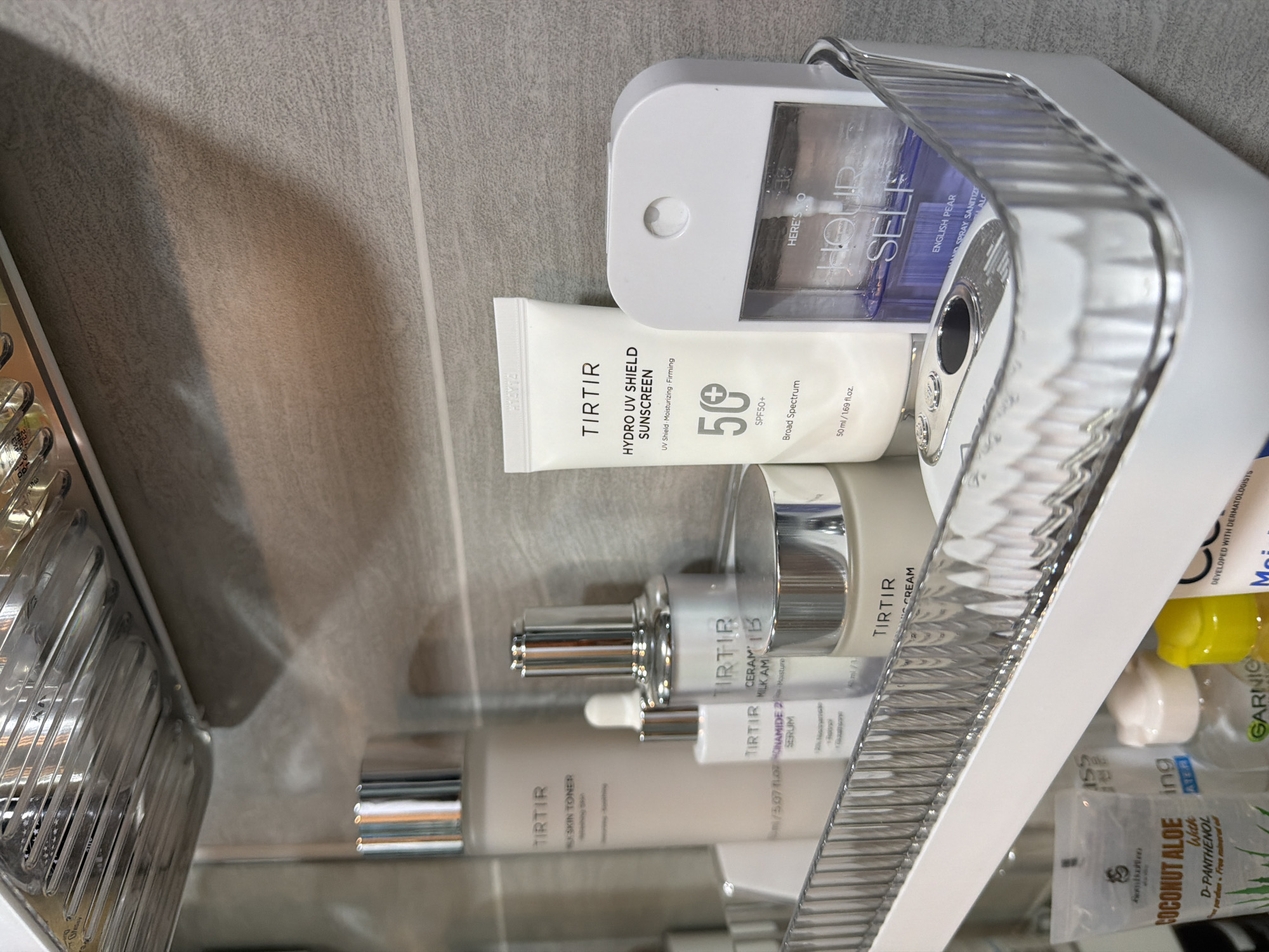 The scene depicts a well-organized bathroom shelf. The geographical location, specifically Bangkok, Thailand, cannot be ascertained from the visual information presented.
The upper portion of the image reveals a metallic, possibly chrome, slotted structure, from which a single water droplet is observed hanging. Below this, the wall is covered in light gray, textured tiles, with a horizontal white grout line visible towards the lower middle of the frame.
A white plastic shelf with a clear, ribbed front edge holds several personal care products. From left to right, these include:
1. A tall, light beige cylindrical bottle with a silver cap, labeled "TIRTIR MILK SKIN TONER."
2. A smaller, clear glass dropper bottle with a silver cap, labeled "TIRTIR NIACINAMIDE 2% SERUM."
3. A silver pump bottle with a silver cap, labeled "TIRTIR CERAMIDE MILK AMPOULE."
4. A short, wide, light beige jar with a silver lid, labeled "TIRTIR CERAMIC CREAM."
5. A white squeeze tube with a black cap, labeled "TIRTIR HYDRO UV SHIELD SUNSCREEN SPF 50+ BROAD SPECTRUM."
6. A white, opaque holder with a clear front, containing a clear bottle of purple liquid labeled "HERE'S YOUR HOUR SELF ENGLISH PEAR HAND SPRAY SANITIZER." The holder features a circular indentation at the top.
7. A white electronic device with two silver circular buttons and a central silver-rimmed opening, positioned to the left of the hand sanitizer.
A lower shelf is partially visible at the bottom of the image, holding additional products including a clear pouch labeled "COCONUT ALOE WITH D-PANTHENOL," a clear bottle with a white cap labeled "GARNIER MICELLAR CLEANSING WATER," a yellow-capped bottle, and a blue and white bottle partially visible, labeled "CERAVE."
No individuals are present in the image. No actions or interactions are depicted; the scene is static. The overall environment suggests a modern bathroom setting.