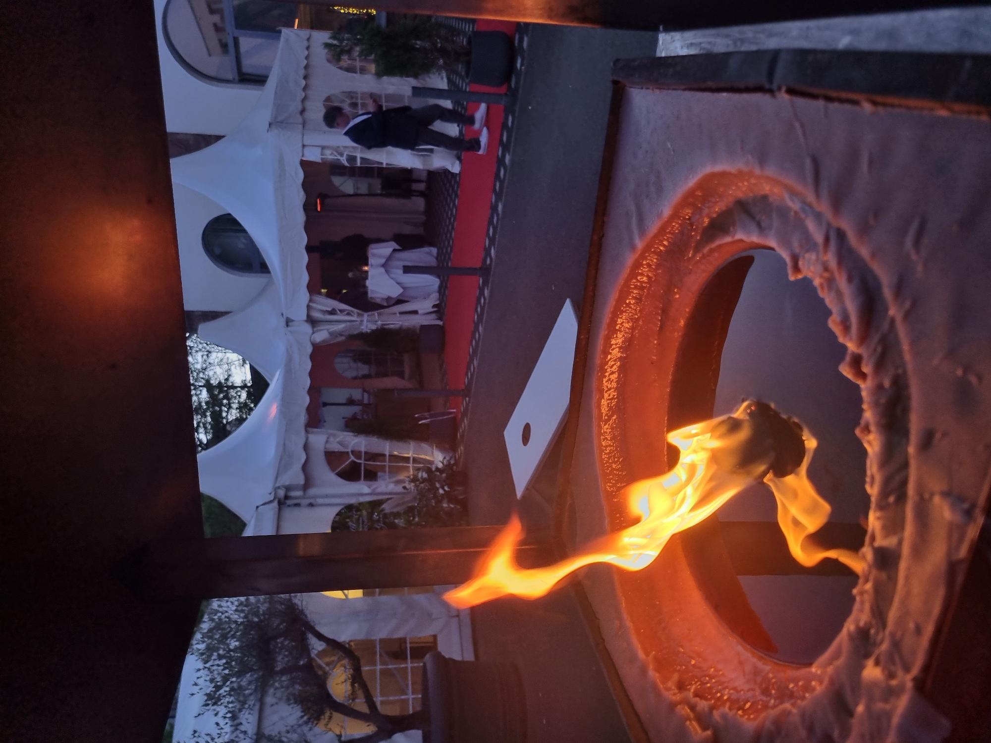 An outdoor evening event is depicted, with a warm fire pit in the foreground and a festive setup behind it. The image is oriented vertically, making the ground appear on the left.
In the immediate foreground, a large, dark outdoor fire pit is prominent on the right. A vibrant, orange-yellow flame burns brightly from its circular opening, illuminating the textured, possibly metal, surface around it.
Behind the fire pit, a red carpet stretches across the mid-ground. An adult male, dressed in dark trousers, a dark jacket, a light shirt, and white shoes, walks on the carpet, facing away from the viewer towards a series of white event tents. To the right of the carpet, a white board with a circular hole, resembling a cornhole game board, rests on the ground.
The background is filled with multiple white, peaked event tents. Some tents feature arched window-like openings, and tables with white tablecloths can be seen inside one tent. Dark trees and foliage are visible around the structures, suggesting a garden or park setting. Ambient lighting, possibly from fairy lights or distant building lights, twinkles in the far background, confirming the evening or nighttime setting. The scene conveys a sophisticated or celebratory atmosphere. No discernible text is visible.