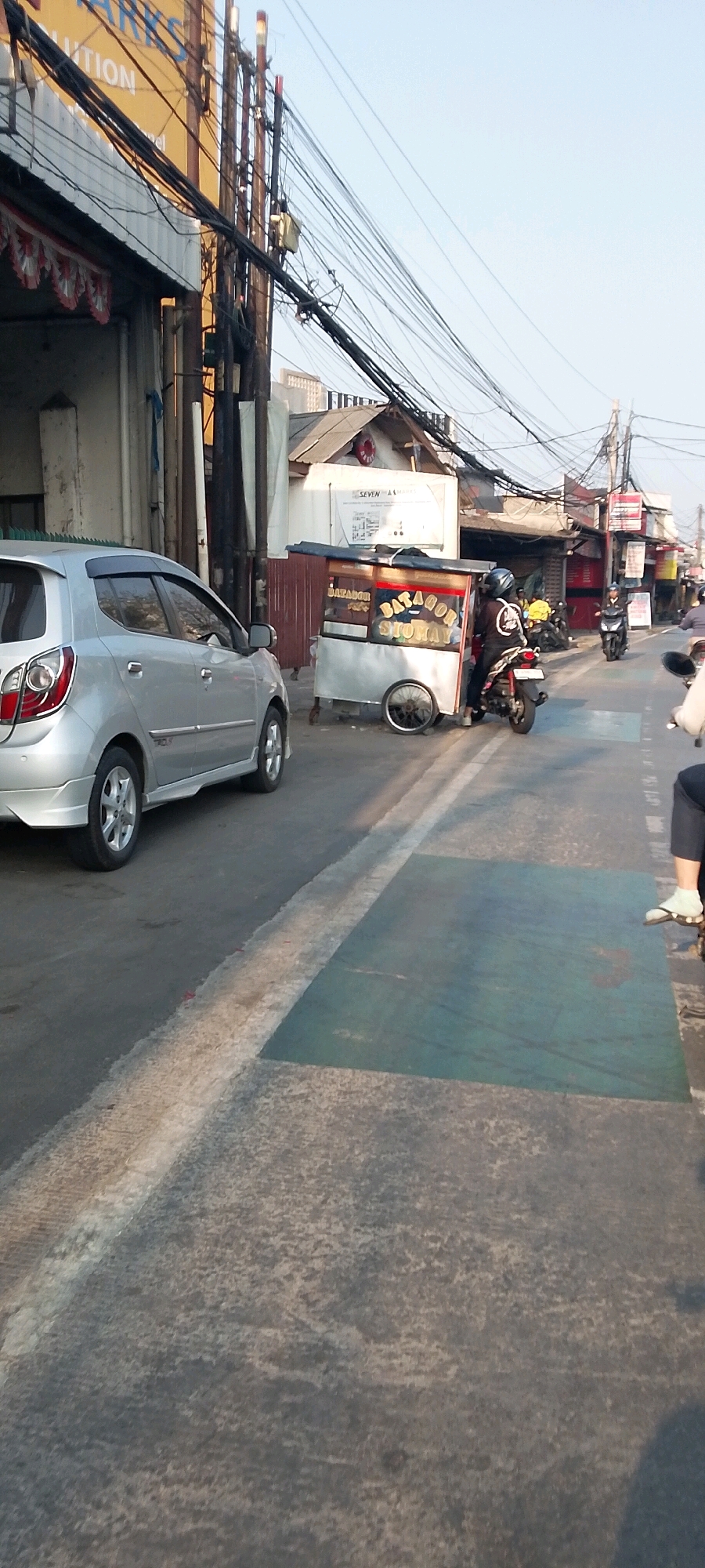 A silver sedan, likely a Toyota, is parked on the left side of the image on a paved road in Bekasi, Indonesia. The car is positioned facing away from the viewer and appears to be a commercial street with various shops and businesses lining the right side of the road. In the background, a food cart selling "BATAGOR SIOKAY" is visible, with a person on a motorcycle near it. The street is cluttered with numerous electrical wires overhead, a common sight in Indonesian urban areas. The sky is clear and bright, suggesting it is daytime. The overall impression is a bustling street scene in a typical Indonesian city.