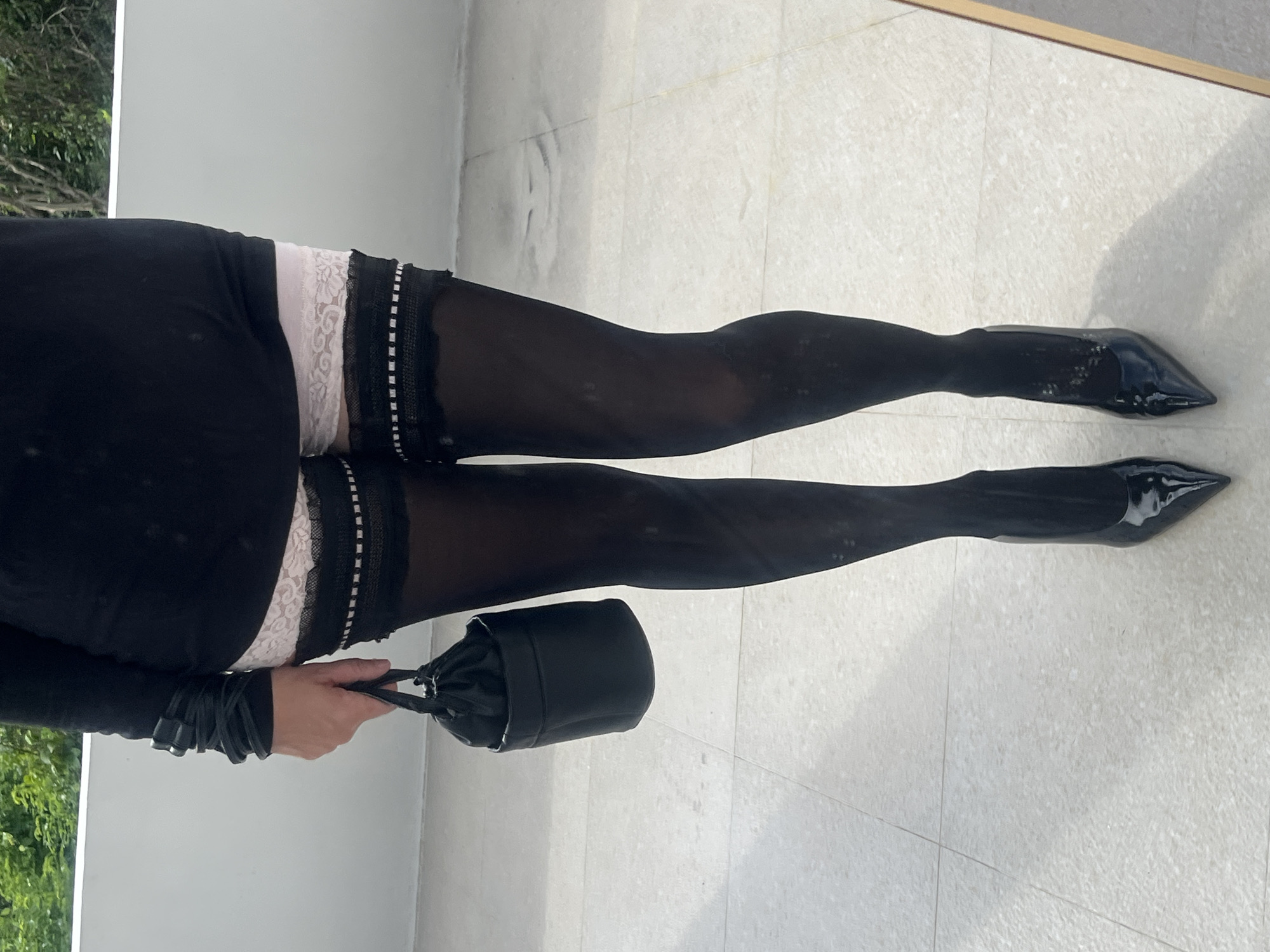 A vertical, full-length shot of a person standing, rotated 90 degrees clockwise from its original orientation. The person's upper body is mostly out of frame, but they are wearing a black long-sleeved top or dress. Below this, a white lace garter with a floral pattern is visible, peeking out just above the knees. Black sheer thigh-high stockings cover the legs, leading down to glossy black pointed-toe high heels.
The person is holding a small black drawstring bucket bag in their left hand, which is positioned at their side. The hand is visible, wearing a black long-sleeved garment that matches the upper attire.
They are standing on a light-colored tiled floor, possibly outdoors or in a brightly lit indoor space, such as a balcony or patio in Kuta Selatan, Indonesia. The tiles are light grey or off-white with visible grout lines. A white wall or railing forms the background, with green foliage visible on the far left, suggesting an outdoor setting. The bright, natural lighting indicates it is daytime, casting a faint shadow on the floor. No text is visible in the image.