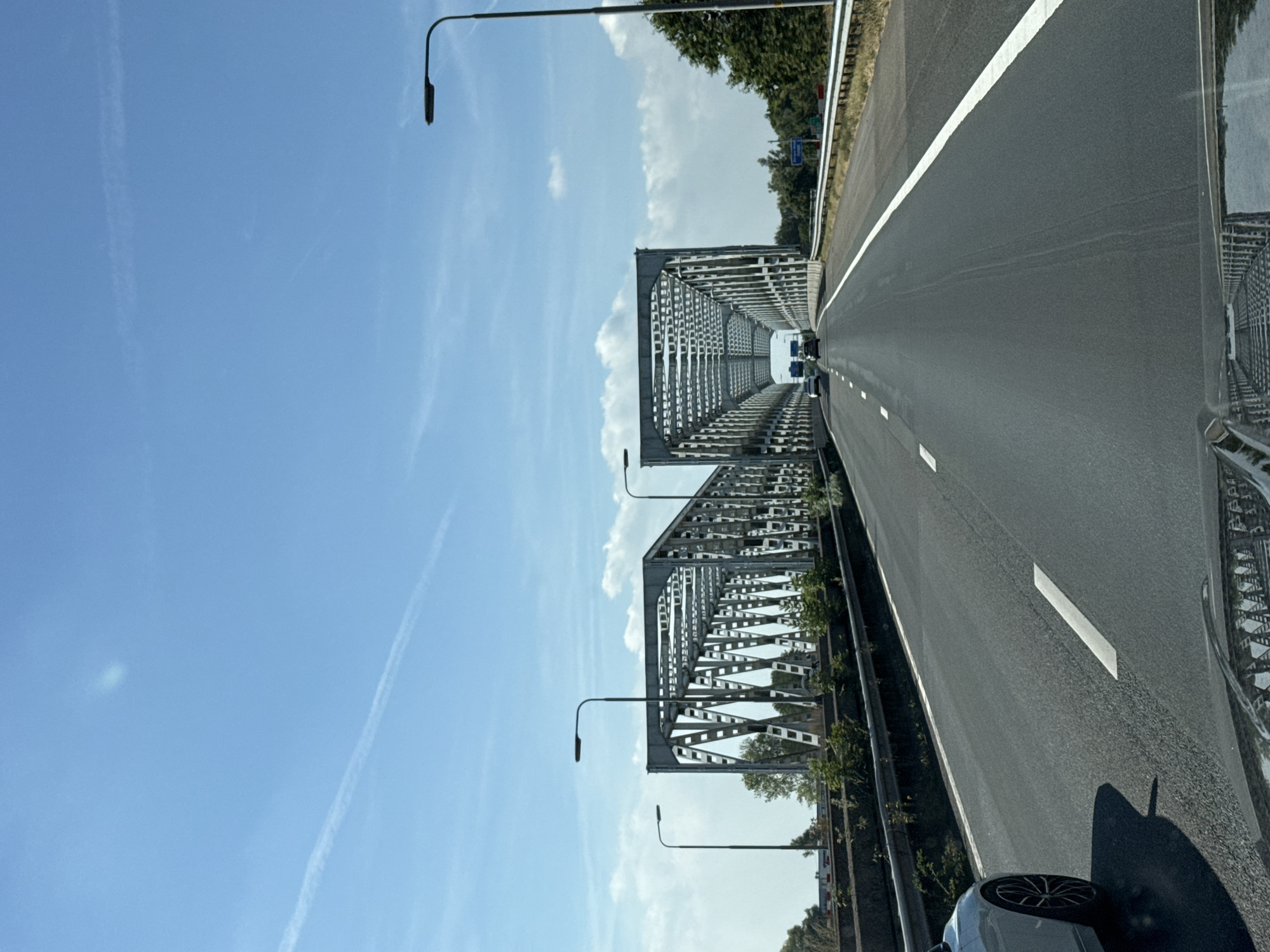 An eye-level, medium shot captures a perspective from a vehicle traveling on a highway towards a large, gray, truss bridge under a clear blue sky with a few wispy white clouds and a faint contrail. The road is paved with asphalt and marked with white lane lines. On the left side of the road, a concrete barrier topped with a metal guardrail separates the highway from a raised embankment with sparse vegetation. Several streetlights with dark, angled lamps are visible along the highway.
The bridge structure is prominent, appearing to be composed of two parallel truss spans. The open latticework of the bridge is a repeating pattern of triangles and rectangles. In the distance, other vehicles can be seen on the highway as they approach and enter the bridge. The surrounding environment beyond the highway appears to be a mix of trees and vegetation. The overall scene suggests a sunny day, possibly in a rural or suburban area. The bottom of the image shows a reflection of the bridge structure in what appears to be a car's windshield or dashboard.