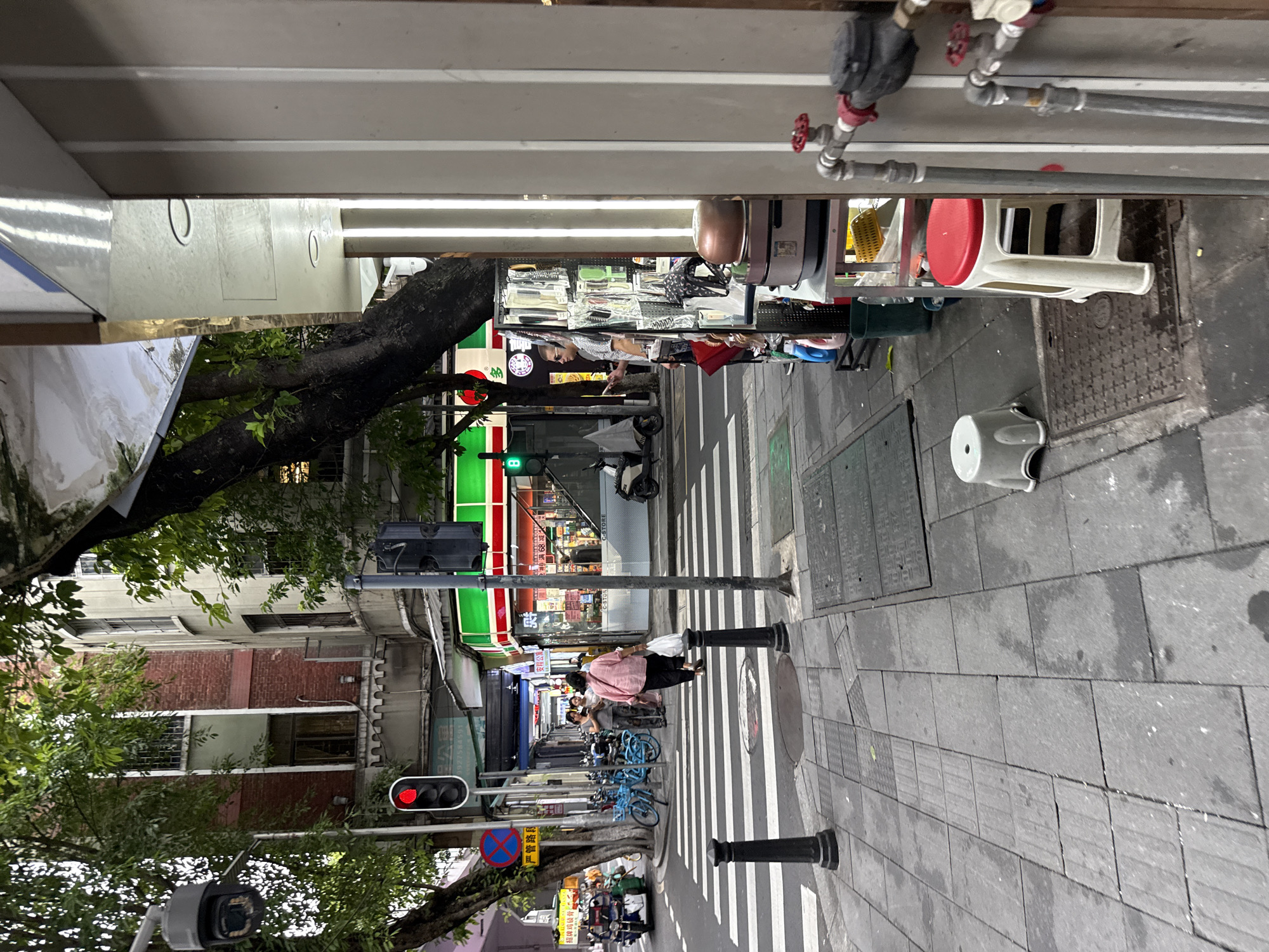 This image depicts a street scene in Yuexiu District, China. A red traffic light is visible on the left, with a pedestrian crossing in the foreground. Several people are present, including a woman in a pink top carrying a white plastic bag, walking across the street. To the right, a man wearing sunglasses stands in front of a display of items, possibly for sale, next to a store with a green and red awning. Bicycles are parked along the sidewalk on the left. The environment appears to be urban, with buildings and trees lining the street.