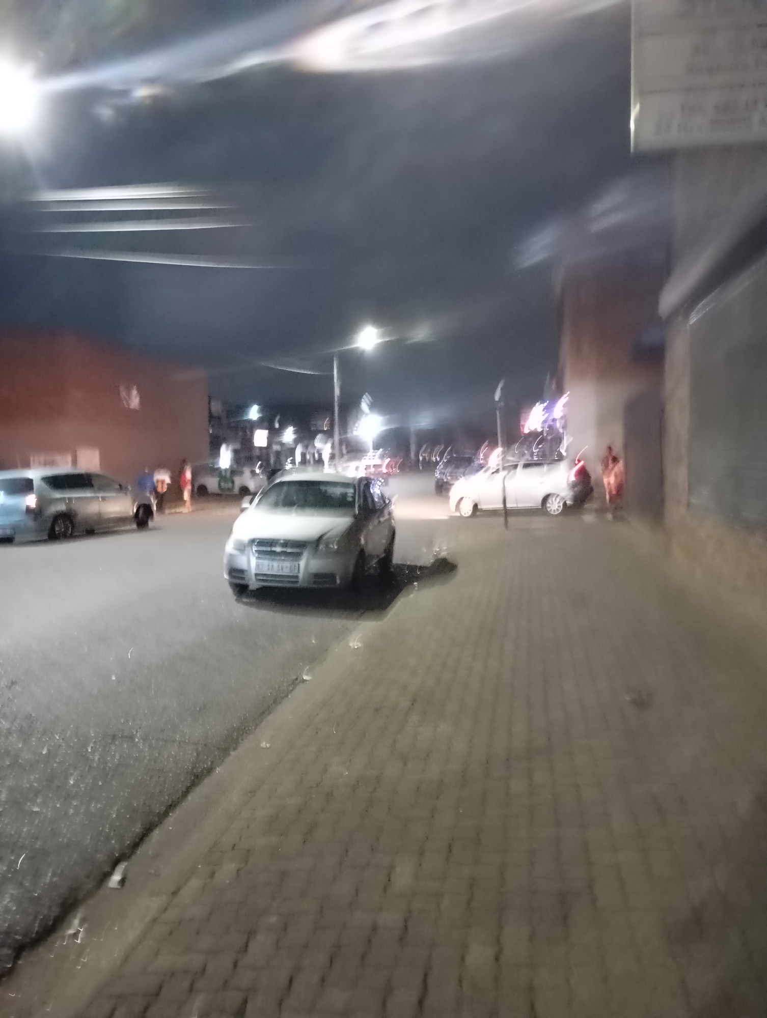 The image shows a street scene at night in what appears to be a residential or commercial area in Johannesburg, South Africa. Several cars are parked along the side of the road, with one silver-colored car prominently in the foreground. There are people walking along the sidewalk. The lighting is provided by street lamps, and the overall ambiance is somewhat blurry, suggesting the photo may have been taken while in motion or with a less-than-stable camera. Buildings are visible on either side of the street, with some illuminated signs or decorations in the background. The road surface appears to be asphalt, while the sidewalk is paved with patterned bricks.