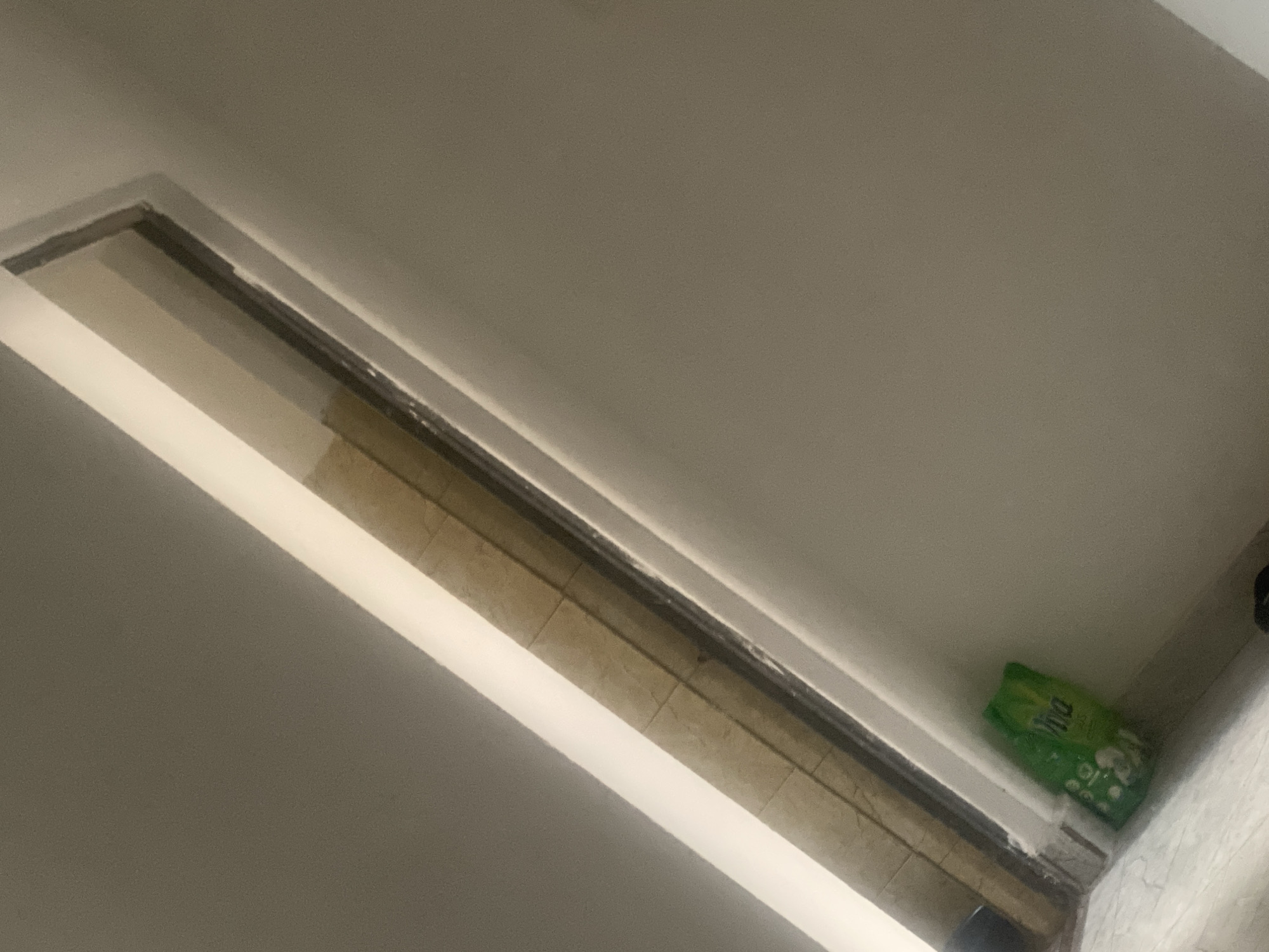 The image displays an interior architectural detail, likely within a building in Jalingo, Nigeria, although no visual cues in the image directly confirm this specific geographical location. The scene is dominated by a long, narrow, rectangular aperture or recess, positioned diagonally across the frame from the upper left to the lower right.
The inner surface of this aperture is lined with small, rectangular, light brown or beige tiles, arranged in horizontal courses. These tiles feature visible grout lines, suggesting a grid pattern. The aperture is framed by smooth, white or off-white painted surfaces, possibly plasterwork or trim, which define its edges against a lighter, neutral-colored wall or ceiling. A dark, thin line runs along the immediate inner edge of the white framing, potentially indicating a shadow, a sealant, or a dark interior frame component.
In the bottom-right portion of the image, resting on a light-colored tiled floor with a subtle pattern, is a green plastic bag or package. The word "Viva" is partially visible on the bag, along with other unidentifiable white markings. The surrounding walls and ceiling appear to be painted in a uniform, light, neutral tone, possibly off-white or light gray. No individuals or specific actions are depicted in the frame. The lighting appears to be interior and somewhat diffused.