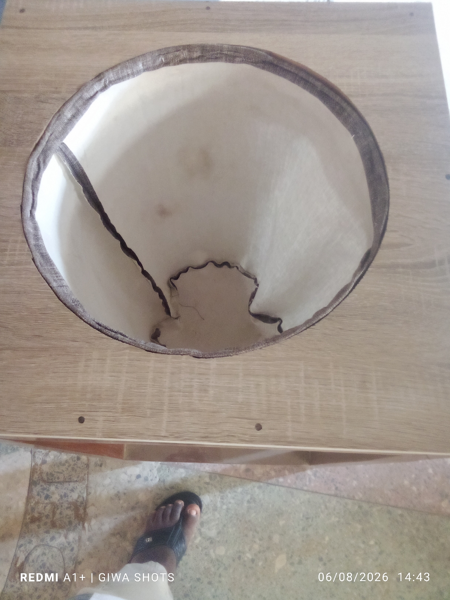 A high-angle, full shot shows a wooden structure with a circular opening lined with fabric. The fabric is light-colored and appears to be a textured material, possibly canvas, with a darker fabric trim around the opening. The trim is frayed in places, and the interior fabric is wrinkled and creased. The wooden surface is light brown with a visible wood grain pattern. Below the wooden structure, a tiled floor is visible, with one foot wearing a sandal positioned on the tiles. The foot appears to be wearing dark-colored sandal with a thong. Text overlays in the bottom left and bottom right corners of the image read "REDMI A1+ | GIWA SHOTS" and "06/08/2026 14:43" respectively, indicating the device and date/time of the photo. The scene appears to be indoors.