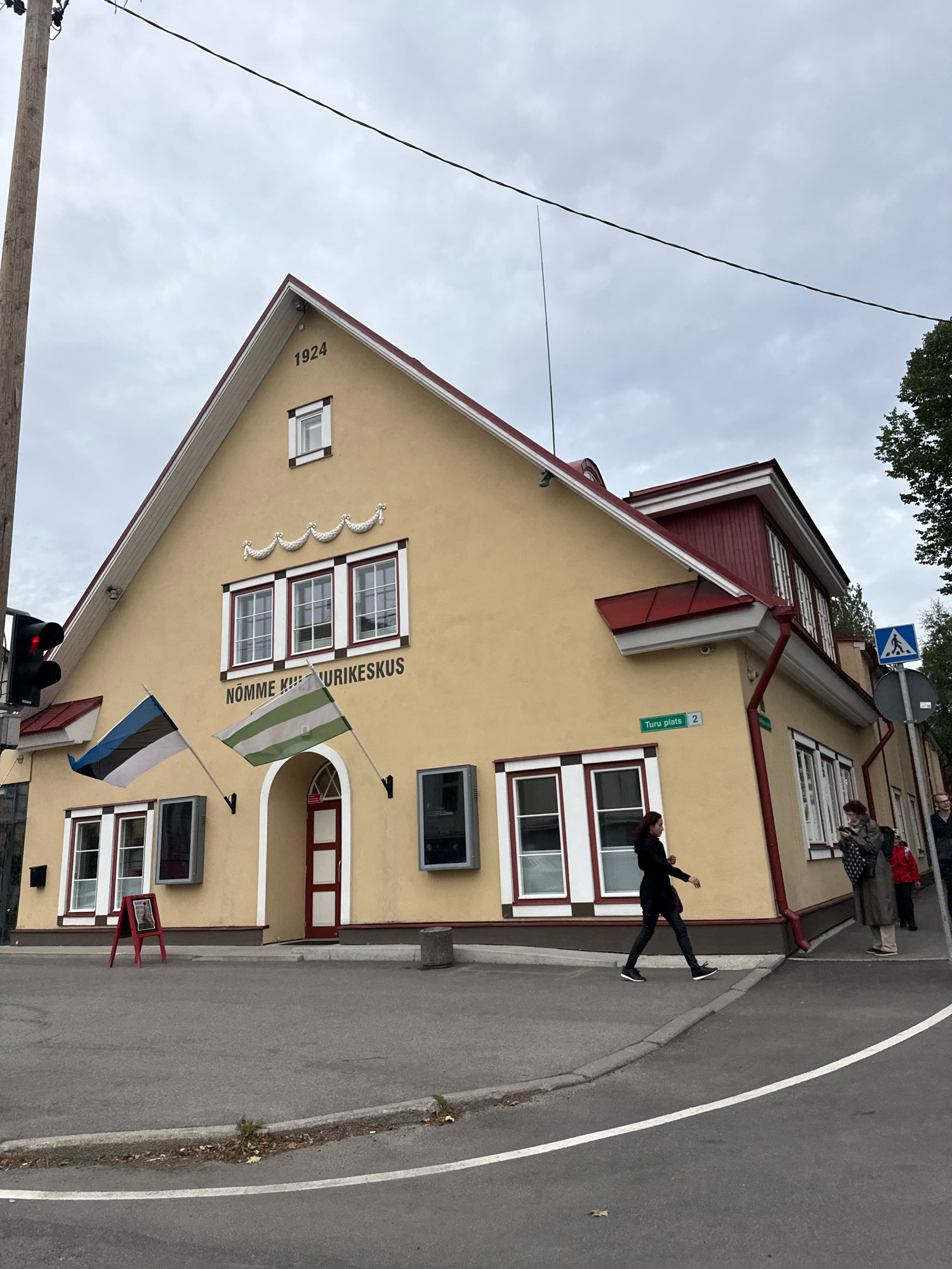 The image shows the Nõmme Cultural Center, located at Turu plats 2 in the Nõmme district of Tallinn, Estonia. The building is a two-story structure with a pale yellow facade and a red roof. The year "1924" is inscribed above the entrance. Two flags are flying in front of the building: the flag of Estonia and another flag with green and white stripes. Several windows are visible, some with white shutters.
In the foreground, a person is walking across the street. Further to the right, two women are standing near the entrance of the building, and another person is visible in the background. A traffic light is on the left side of the image, and a pedestrian crossing sign is on the right. The sky is overcast. The overall scene depicts a street view of a cultural center in an urban setting.