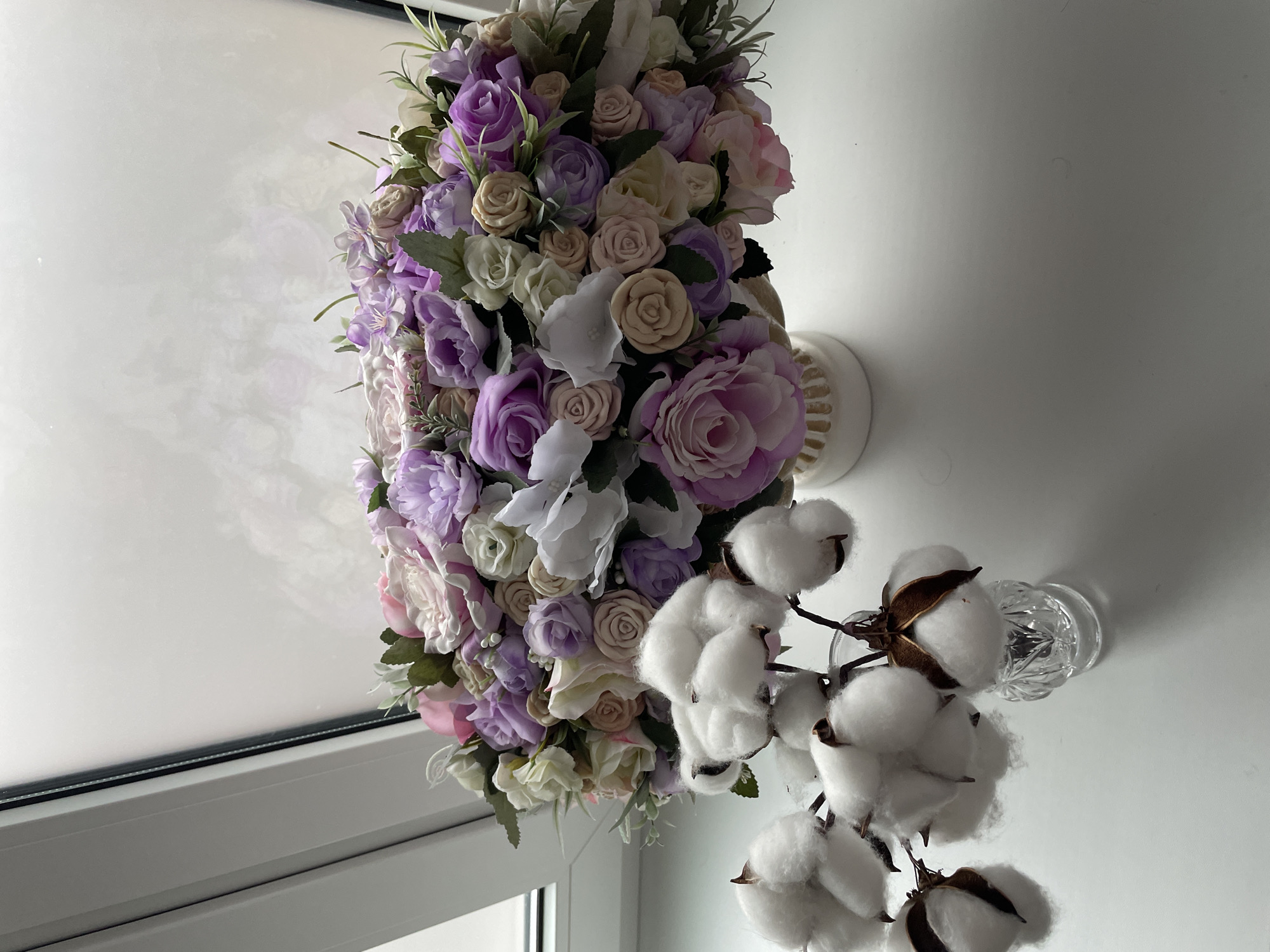 The image shows a bouquet of flowers in shades of purple, pink, and white, positioned near a window. Next to the bouquet is a clear glass vase holding a branch of cotton flowers. The background is a white wall. The image appears to be taken indoors in a bright, possibly residential, setting.