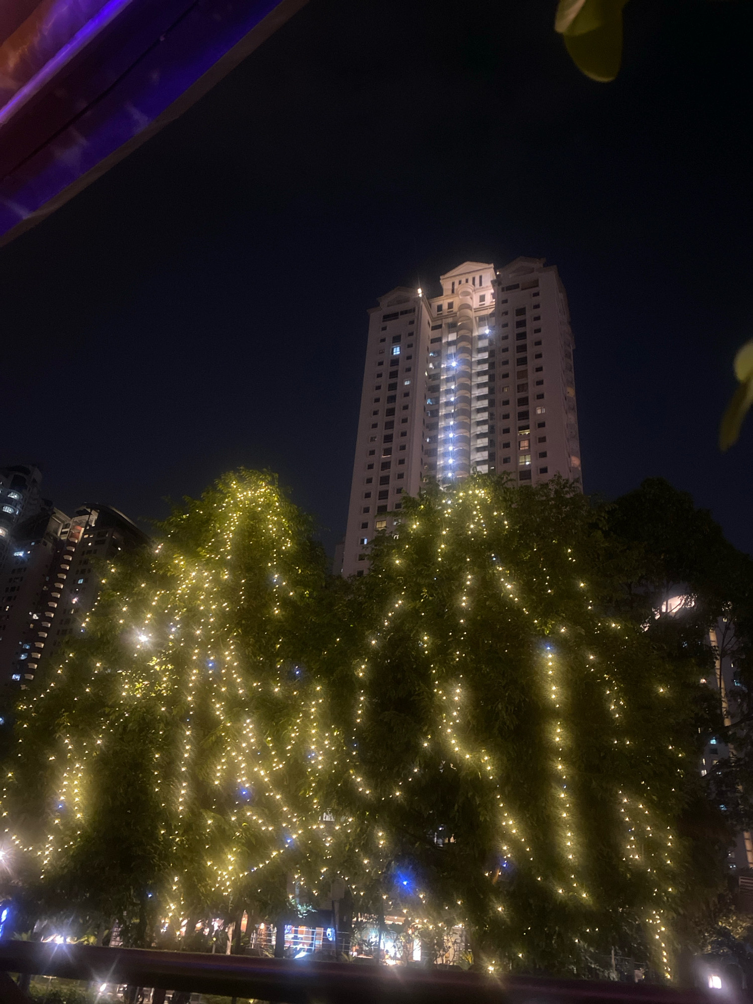 This is a nighttime, low-angle, outdoor shot featuring trees adorned with yellow string lights in the foreground. In the background, a tall, modern apartment building is visible, illuminated by internal lights. The sky is dark, indicating late evening or night. The scene appears to be in a city environment, likely with urban landscaping. The overall impression is of a festive or decorated urban setting. The distinct architecture of the building, particularly its stepped crown, suggests it might be a landmark or residential complex in Kuala Lumpur, Malaysia.