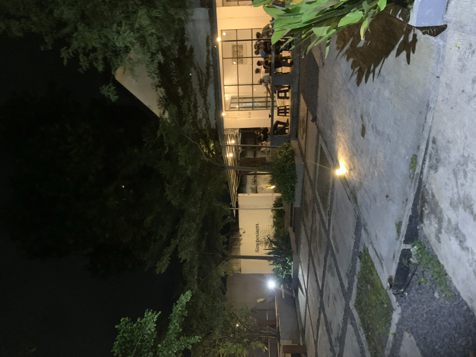 The image shows the exterior of a restaurant or cafe at night, located at the coordinates -7.751, 113.213 in the city of Probolinggo, Indonesia. The building has a white exterior and large windows, with a patio area in front. The patio is made of concrete and is illuminated by several lights. There are a few people sitting at tables on the patio, and some plants are growing in pots. The sign above the entrance reads "Simposium," which suggests that it is a cafe or restaurant. The sky is dark, and the only light source is coming from the building and the lights on the patio. There are trees in the background and the overall ambience is peaceful and relaxing.