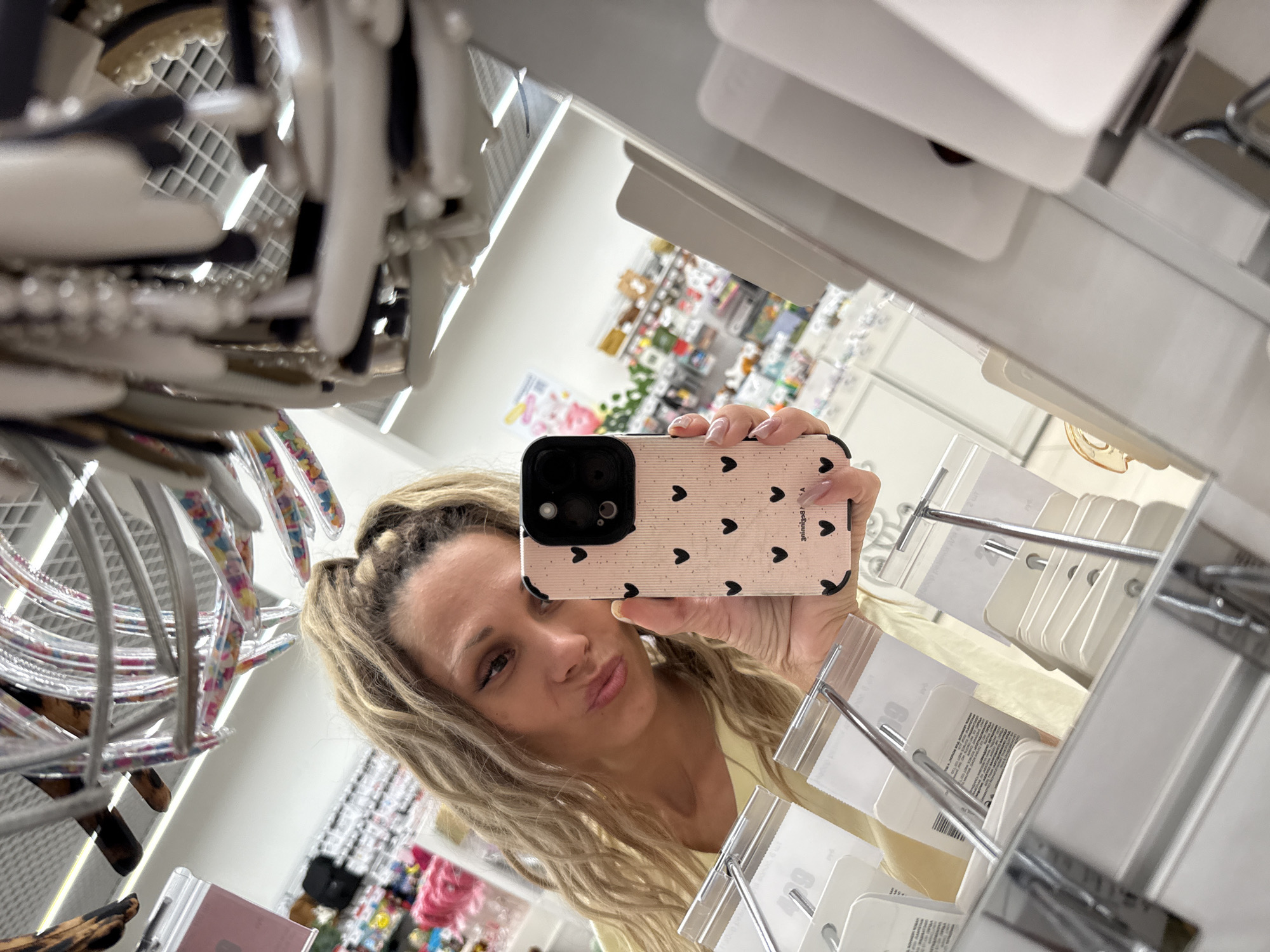 In a retail environment, a woman with blonde, wavy hair pulled into a high ponytail is taking a mirror selfie. She is wearing a light yellow top and is holding a pink smartphone with a black heart pattern in her right hand. The phone case has a camera module with multiple lenses. The woman is making a pouty face towards the mirror. In the foreground, a metal rack displays various hair accessories, including headbands with colorful embellishments and black hair clips. Behind her, shelves are stocked with more merchandise, including what appears to be other hair accessories and cosmetic products. The reflection in the mirror shows a brightly lit store interior with aisles of products and displays. The location is not specified in Sosnovtsy, Russia.