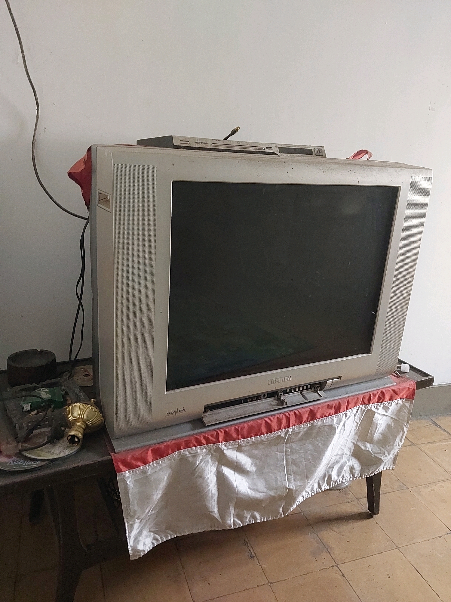 An old, gray Toshiba television with a dark screen sits on a table covered with a white and red cloth. A DVD player is on top of the television. To the left of the television on the table are an ashtray, some CDs, and a golden decorative object. The scene appears to be inside a room with a white wall and tiled floor. The overall impression is one of an older, possibly unused or neglected setup. The image is likely from a residential setting in Malang, Indonesia, given the context of the request.