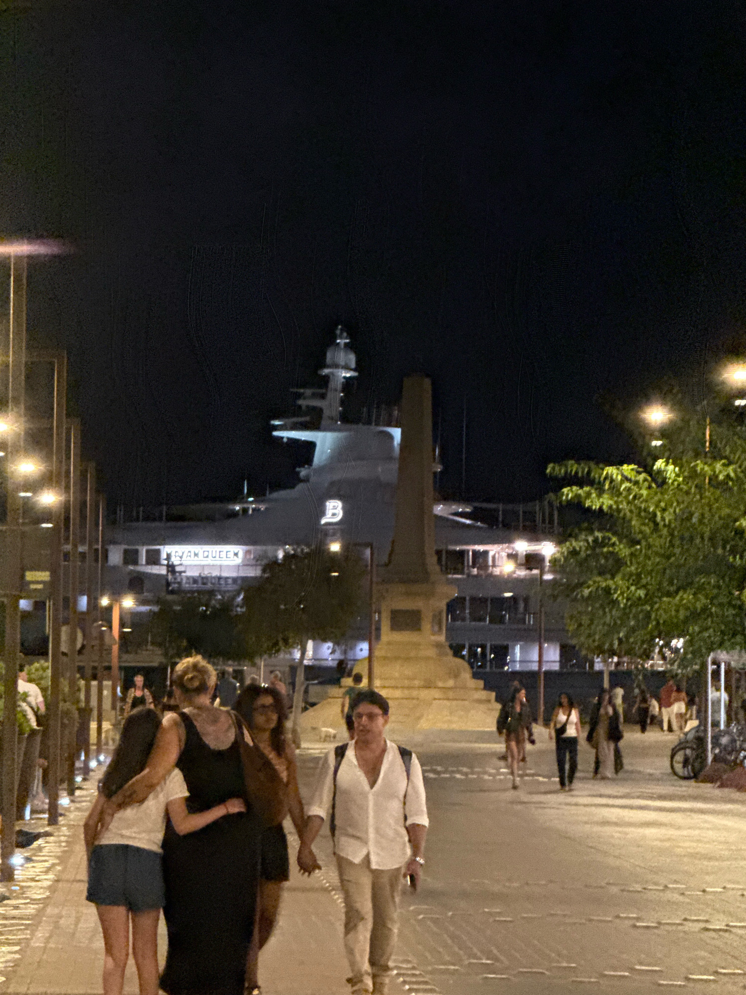 The image depicts a night scene in the city of Ibiza, Spain. In the background, a large white yacht named "MEGAM QUEEN" is docked. In the foreground, a group of people are walking on a paved walkway lined with streetlights. Some of the individuals are walking together, with one woman in a black dress embracing two other women. Another man in a white shirt and light-colored pants is walking in the opposite direction. A monument or obelisk stands to the left of the yacht, and a tree with green foliage is visible to the right. The overall atmosphere is that of a bustling urban environment at night.