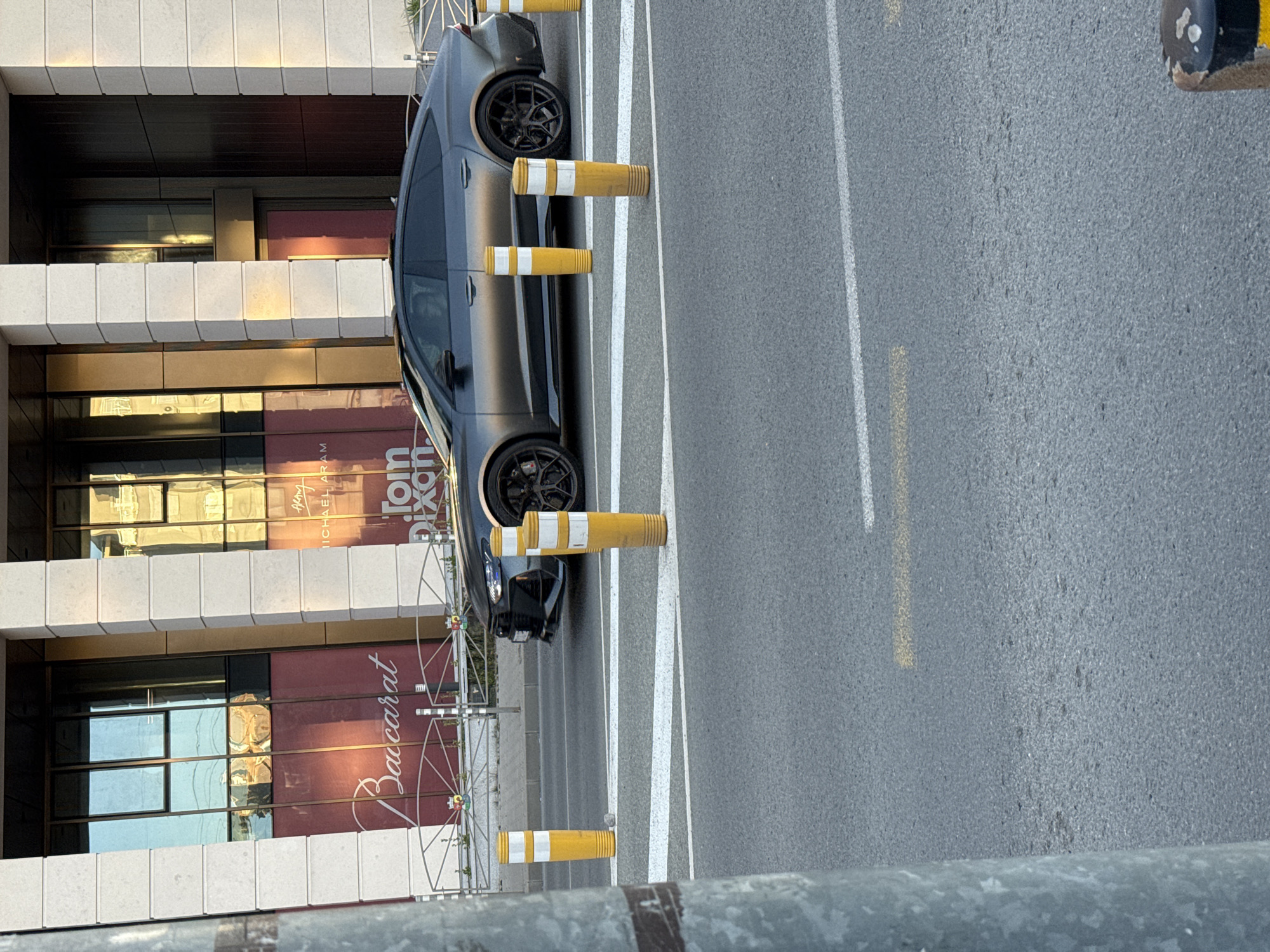 The image shows a dark gray car parked parallel to a building in Sofia, Bulgaria. The building features a sign for "Baccarat" and "Tom Dixon". The car is parked in a lane delineated by white lines and separated from the building by yellow and white striped bollards. The road surface is gray asphalt. The photo is taken from an elevated perspective, looking down onto the scene.