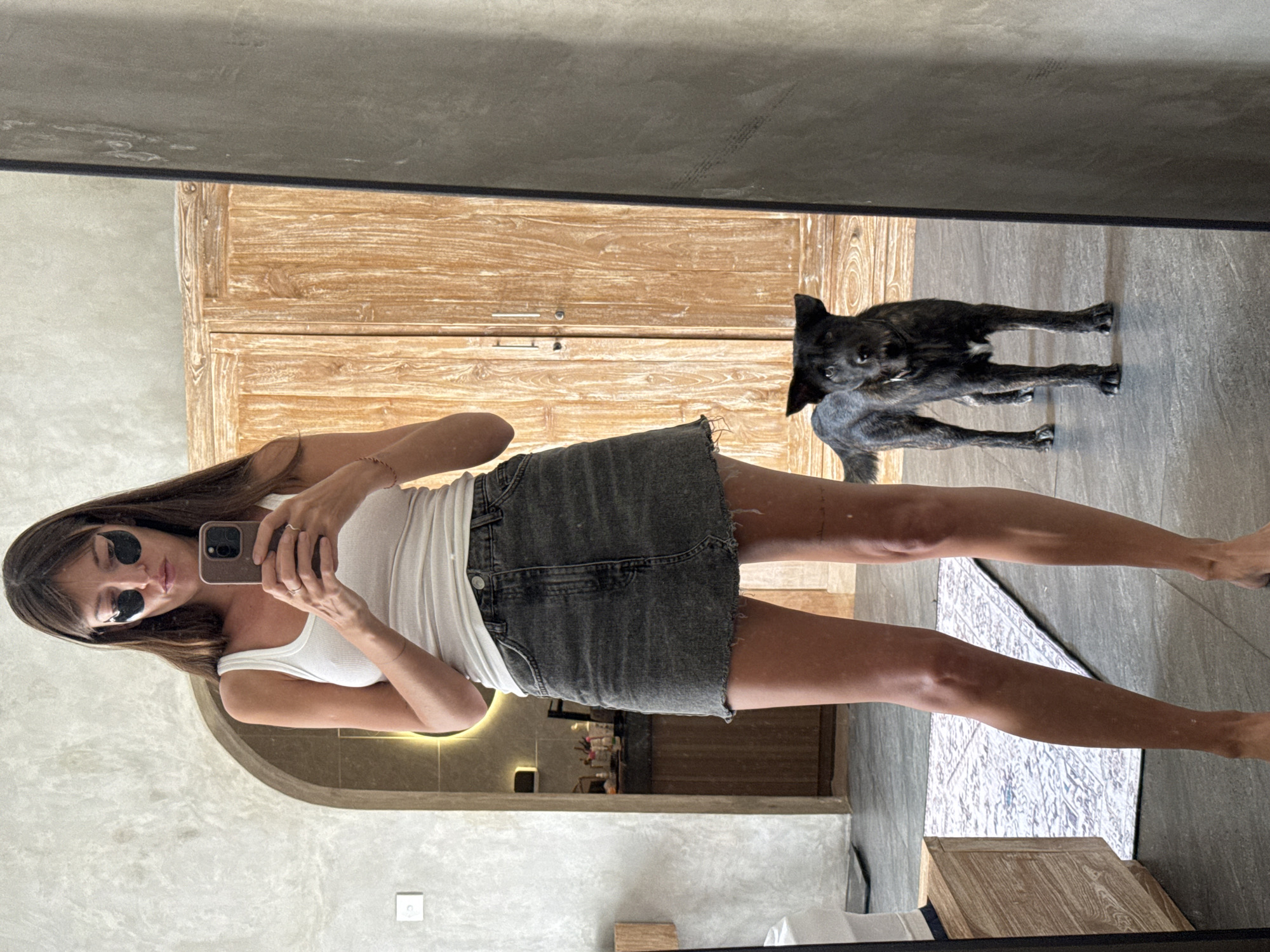 A woman with dark hair and under-eye patches stands in front of a mirror, taking a selfie with her phone. She is wearing a white tank top and a dark denim skirt. A brindle-colored dog stands behind her, reflected in the mirror. In the background, there is a wooden wardrobe, an arched doorway leading to a bathroom, and a patterned rug on the floor. The scene appears to be set in a modern, residential interior, likely a bedroom or dressing area, within a hotel or villa in North Kuta, Indonesia.