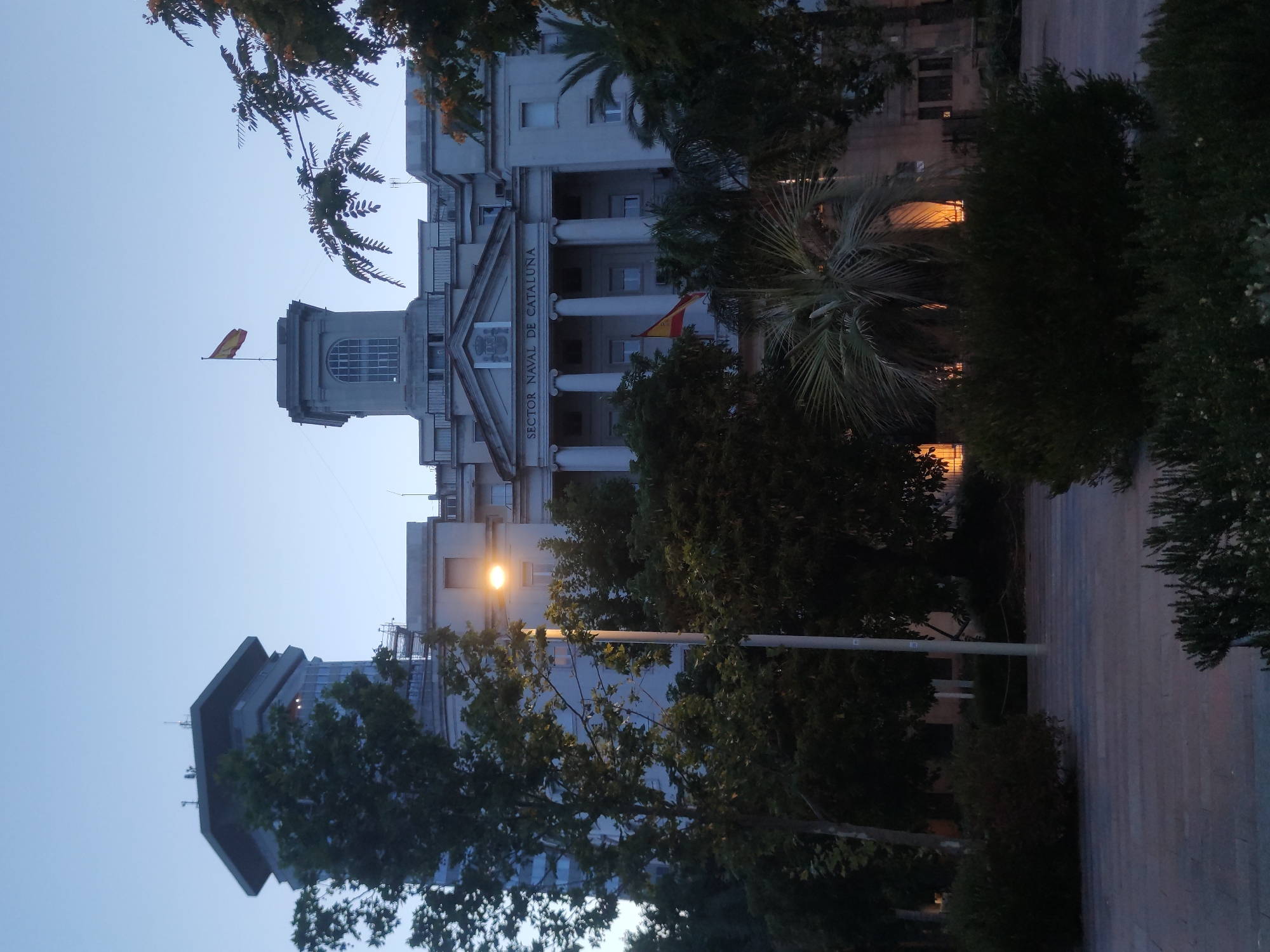The image depicts the building of the Naval Sector of Catalonia in Barcelona, Spain, at dusk. The building is a large, multi-story structure with a neoclassical facade, featuring columns and a pediment above the entrance. The words "SECTOR NAVAL DE CATALUNA" are visible above the columns. A Spanish flag flies from a flagpole on top of the building. To the left of the main building is a taller, more modern tower. The foreground is a paved area with trees and shrubs, some of which are illuminated by outdoor lighting. The sky is a pale blue, indicating either dawn or dusk. There are no people visible in the image.