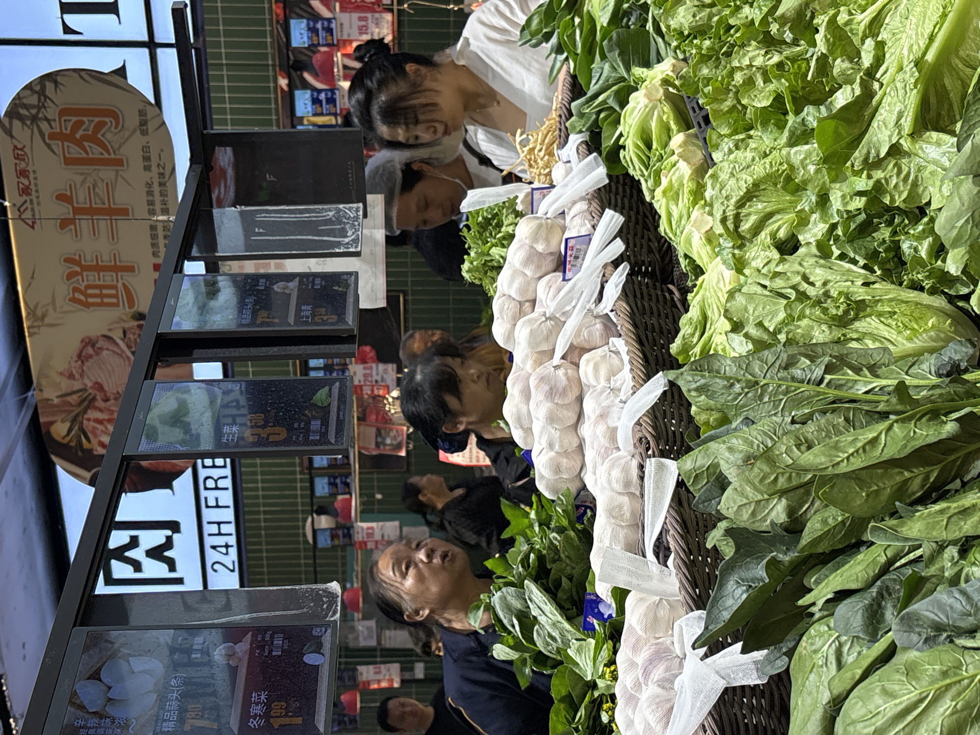 The image shows a vegetable display at a market, likely in Jiangjin, China, based on the language on signs. Several customers are seen browsing the produce, which includes garlic and lettuce. The garlic is bunched together with white netting. Price tags are visible, indicating the cost of different items. The setting appears to be an indoor market.
