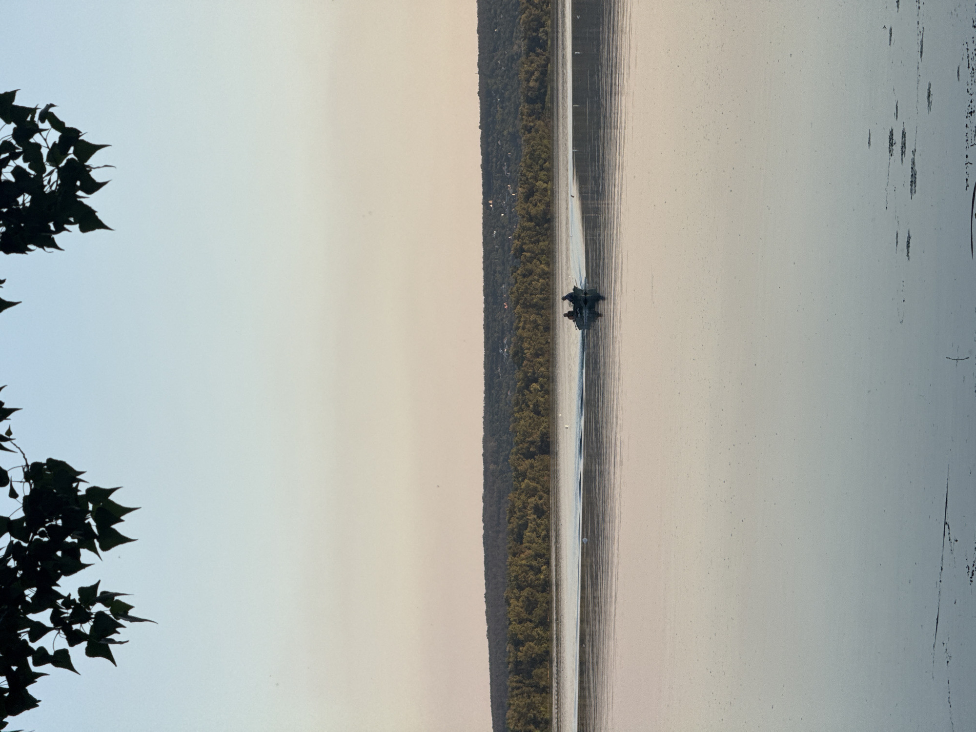 In Obukhivka, Ukraine, on a calm river, two figures are seated in a small motorboat that is moving to the left, creating a wake behind it. The boat is positioned in the middle ground of the image. In the background, a dense line of trees forms a dark, undulating horizon, with a faint scattering of structures visible on the crest of the distant hills. The sky above is a soft gradient of pale blue and pink hues, indicating either early morning or late evening light. In the foreground, the river's surface is smooth and reflective, with dark patches of vegetation or debris visible just below the surface and along the bottom edge of the frame. The top of the image is partially obscured by the dark silhouette of tree branches and leaves hanging from above.